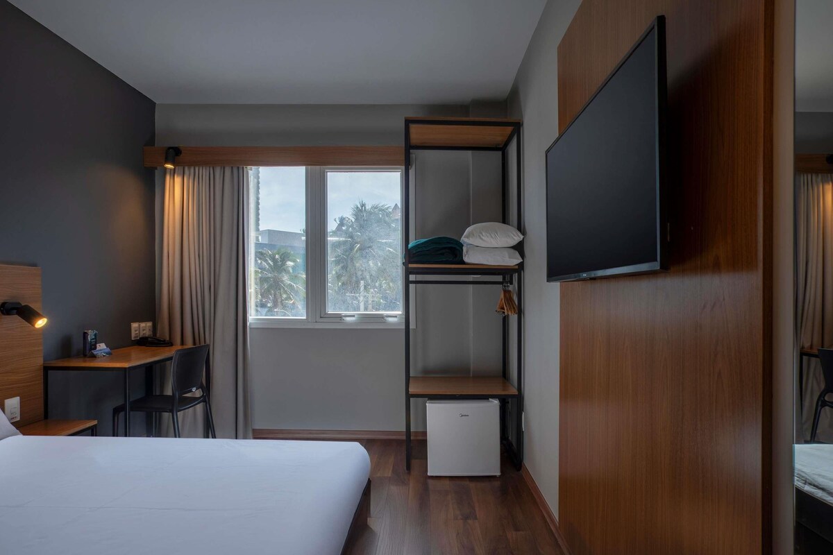 Ein Hotelzimmer mit Bett, Schreibtisch, Stuhl, Fernseher, Kleiderschrank und kleinem Kühlschrank.