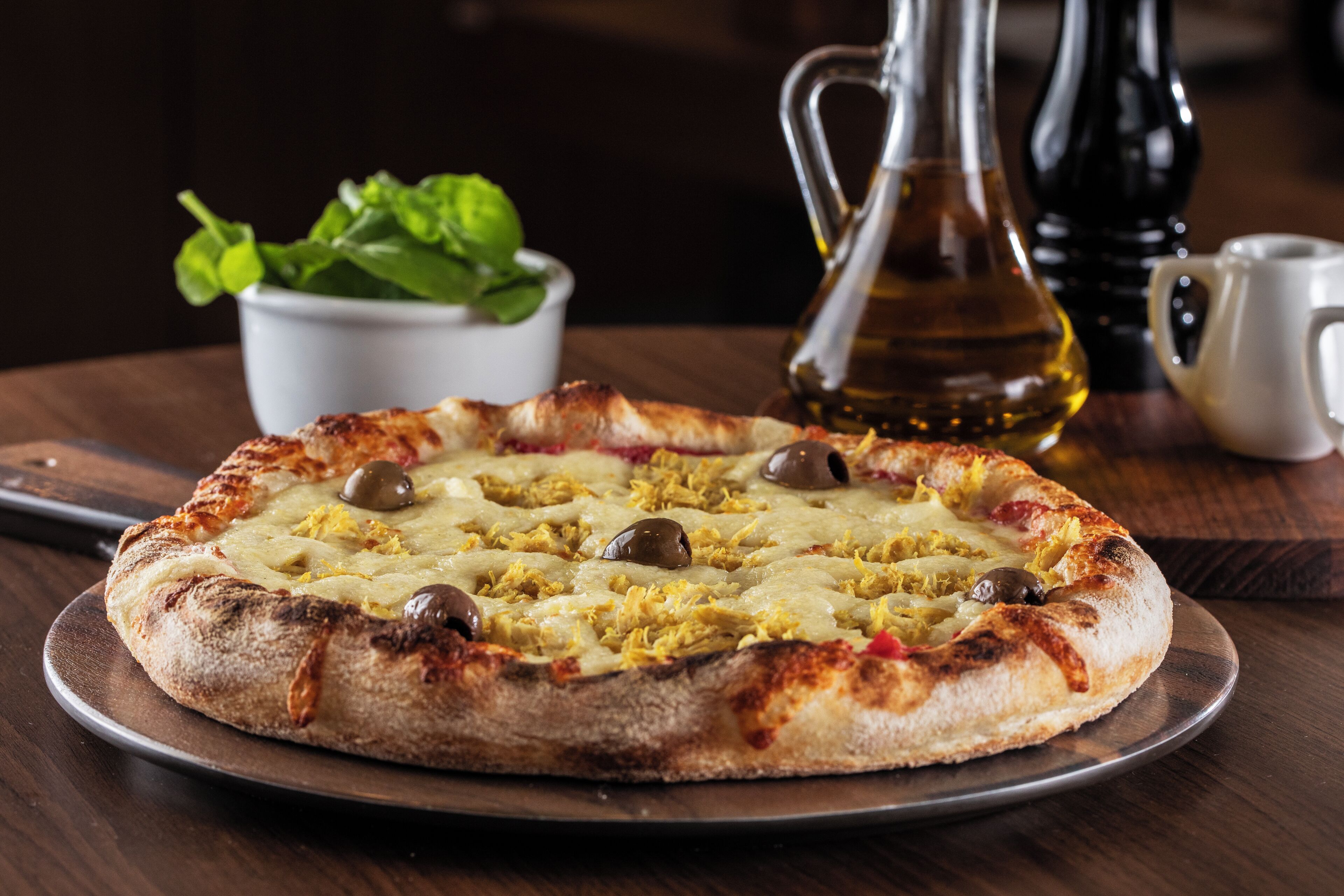 Una pizza sobre una tabla de madera con aceitunas y queso, una jarras de aceite y una taza en una mesa.