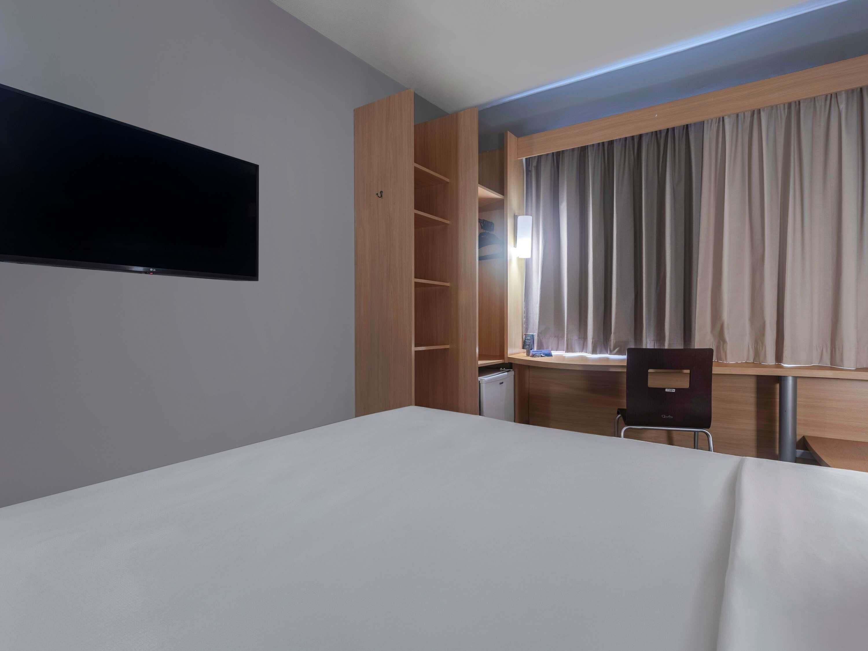 Quarto de hotel com cama, escrivaninha, cadeira, televisão, guarda-roupa e janela com cortinas.