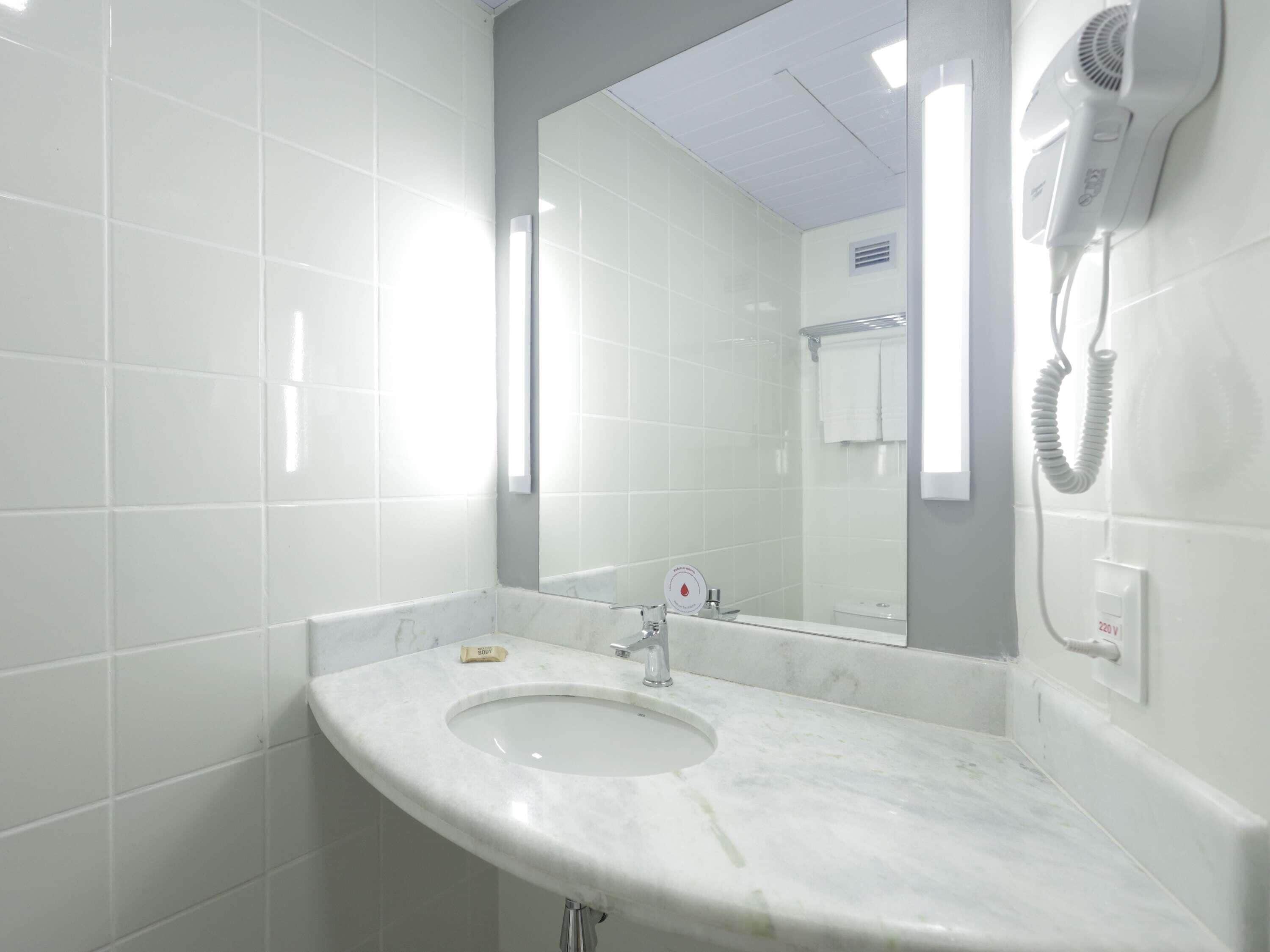 Une salle de bain avec un lavabo blanc, un miroir et un sèche-cheveux fixé au mur.