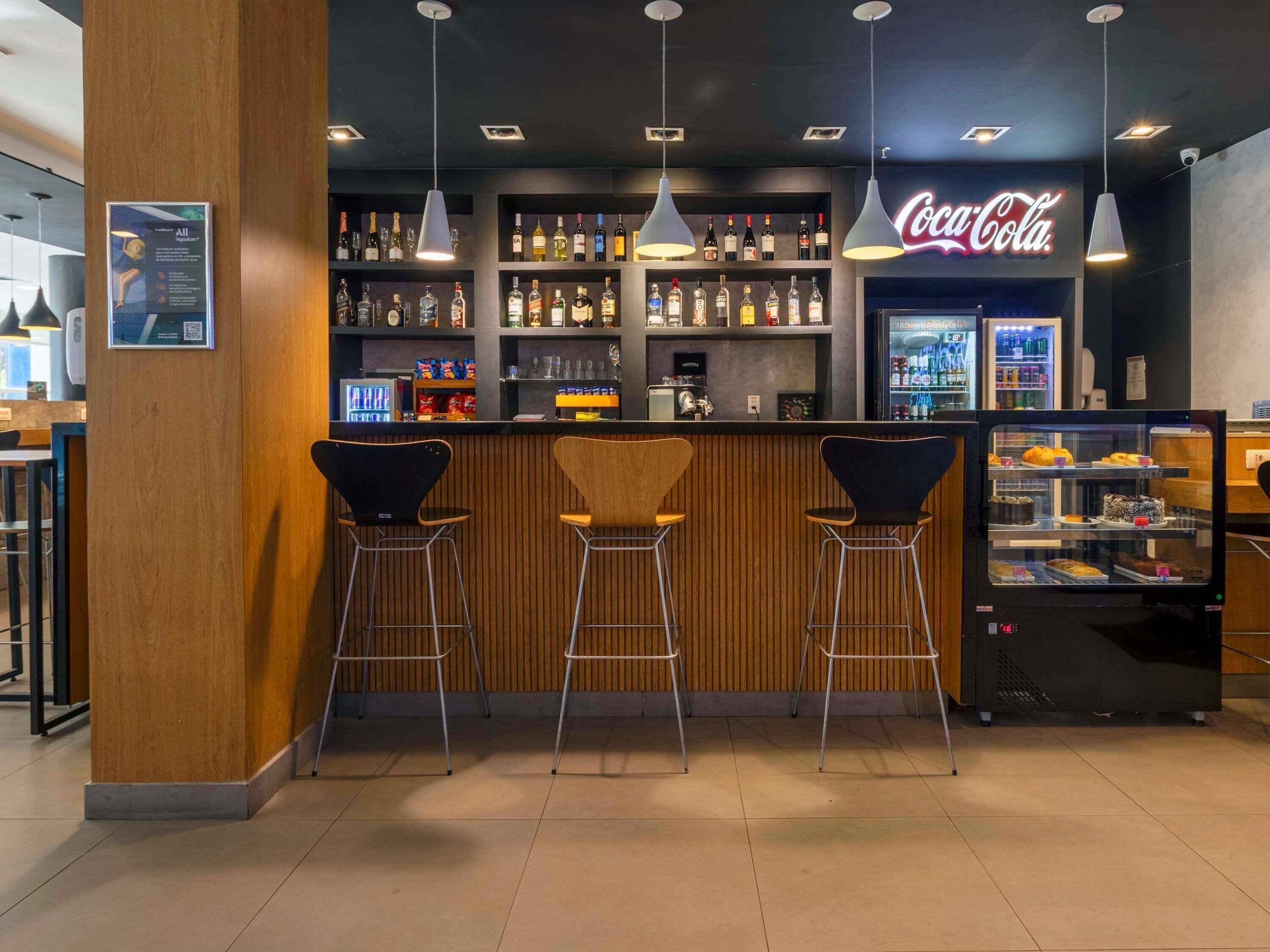 A bar area with three stools, shelves of liquor bottles, a Coca-Cola sign, and a dessert display case.