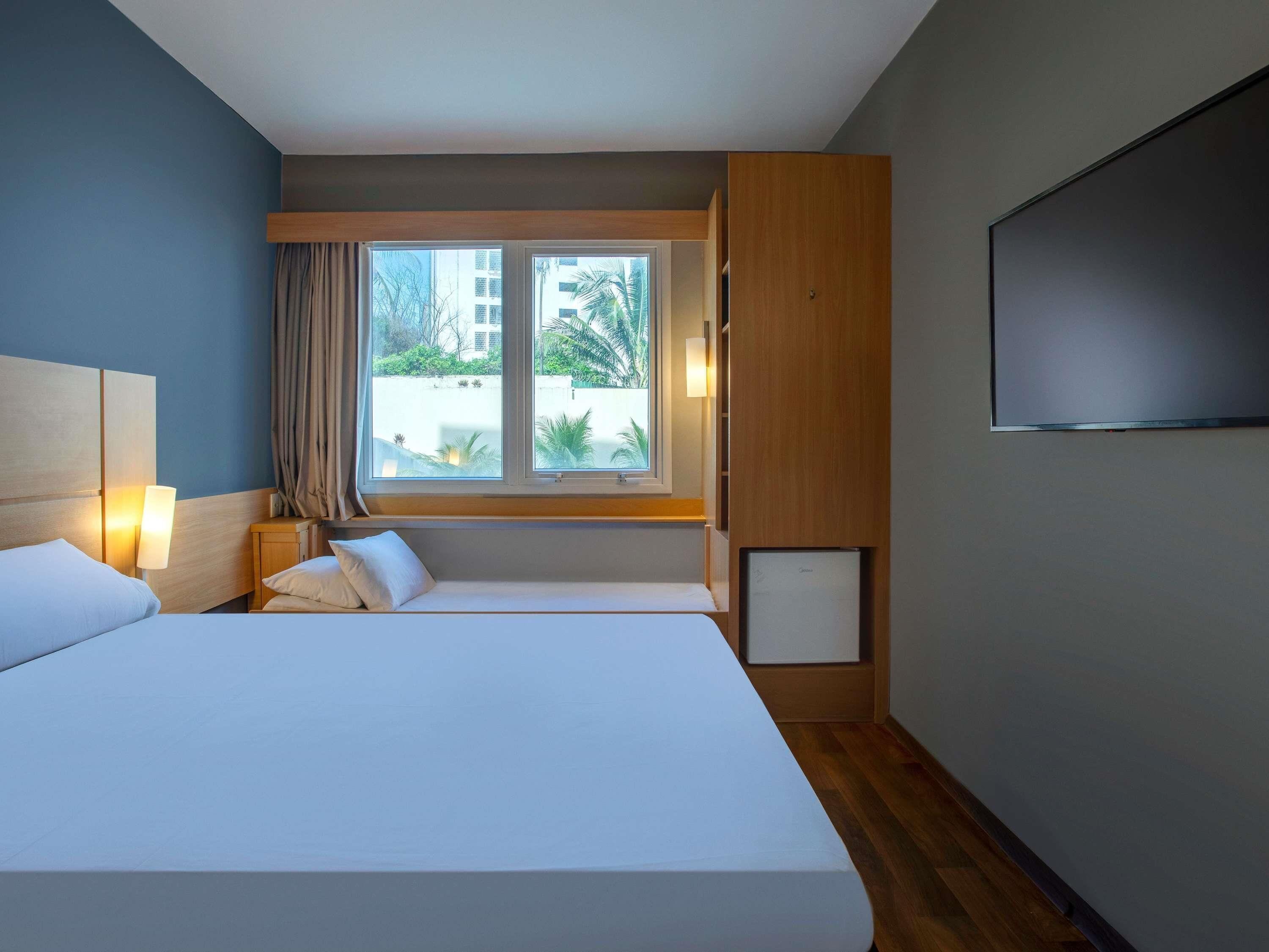 A modern hotel room with a large bed, a TV, a window with a view of buildings, and a wooden cabinet.