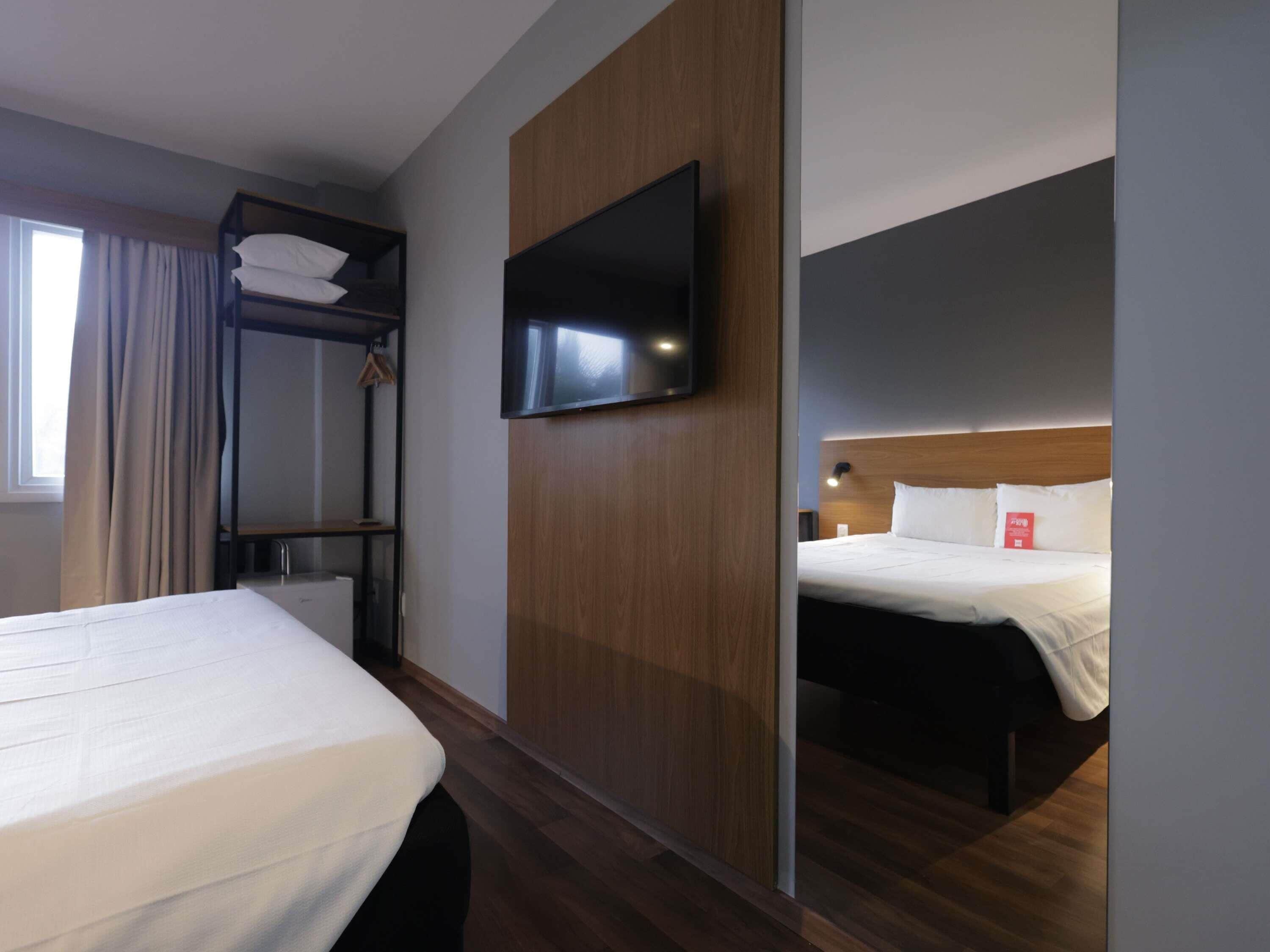 Une chambre d’hôtel équipée d’un lit superposé, d’un lit simple, d’un téléviseur à écran plat fixé sur un panneau en bois et d’une table de chevet avec une lampe.