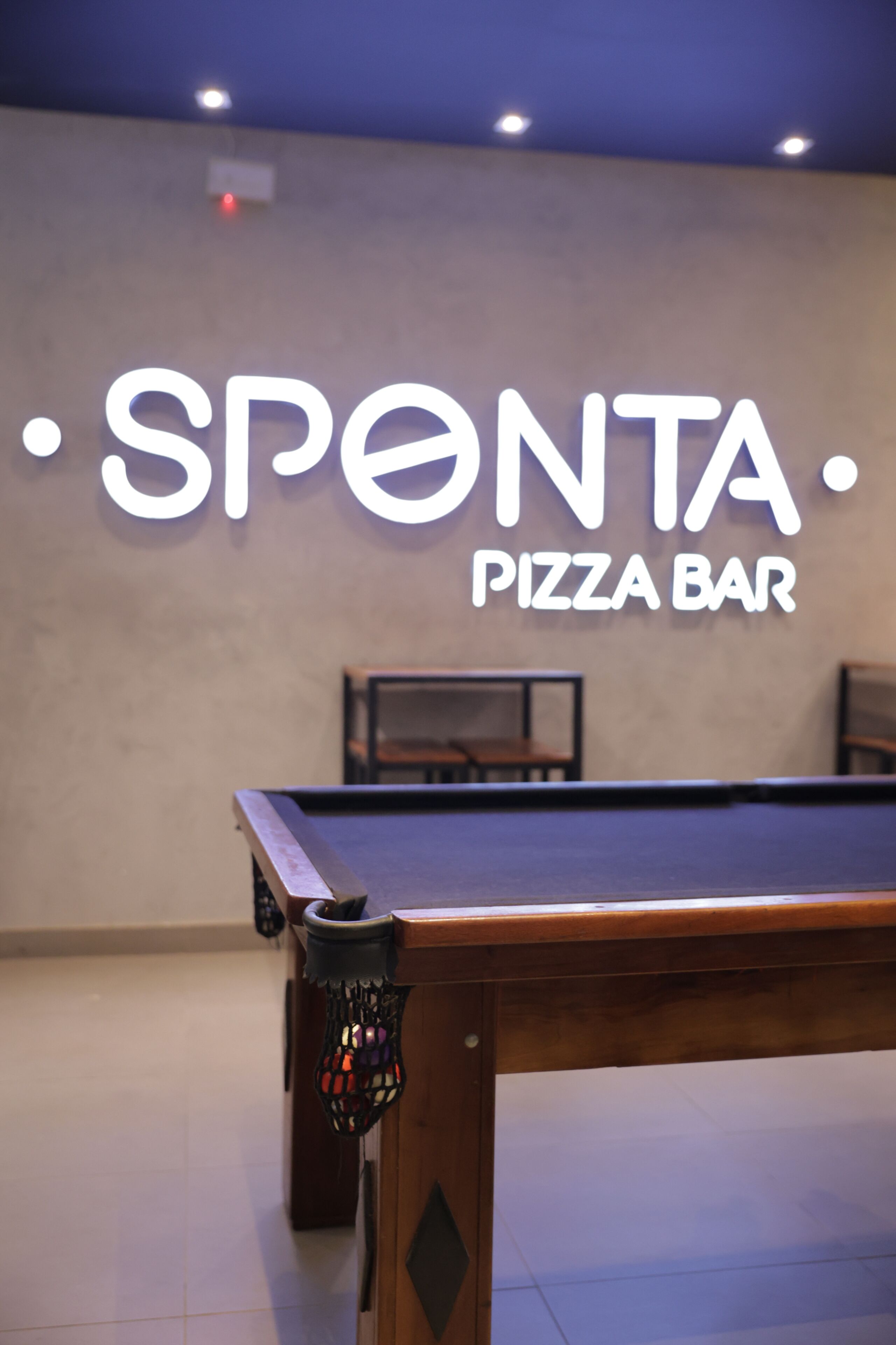 Een biljarttafel met een neonbord voor 'SPONTA PIZZA BAR' op de achtergrond.