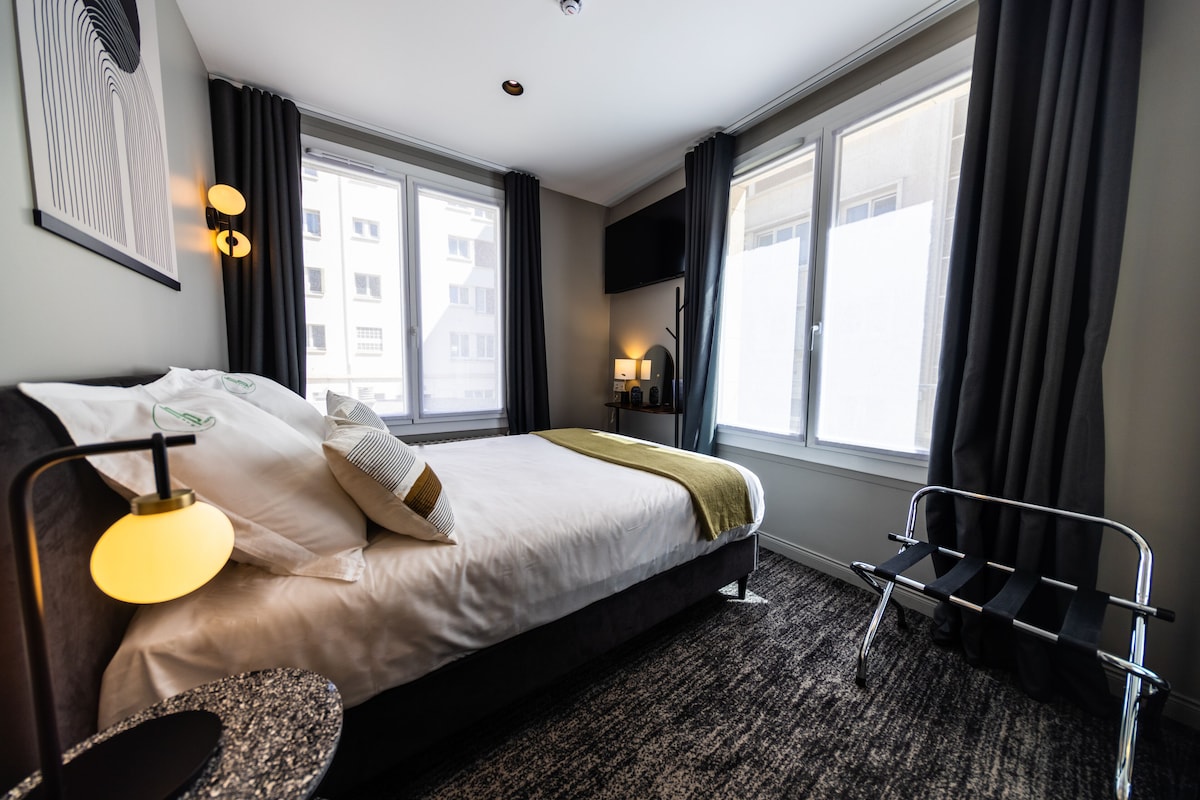 Une chambre d’hôtel moderne avec un grand lit, un bureau et une chaise. Elle est dotée de deux fenêtres avec des rideaux, d’un téléviseur à écran plat et d’une lampe fixée au mur.