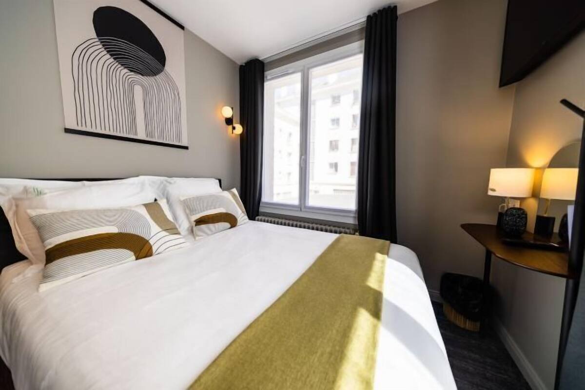 A hotel room with a large bed, a view of buildings through the window, a wall-mounted artwork, and a bedside table with a lamp.