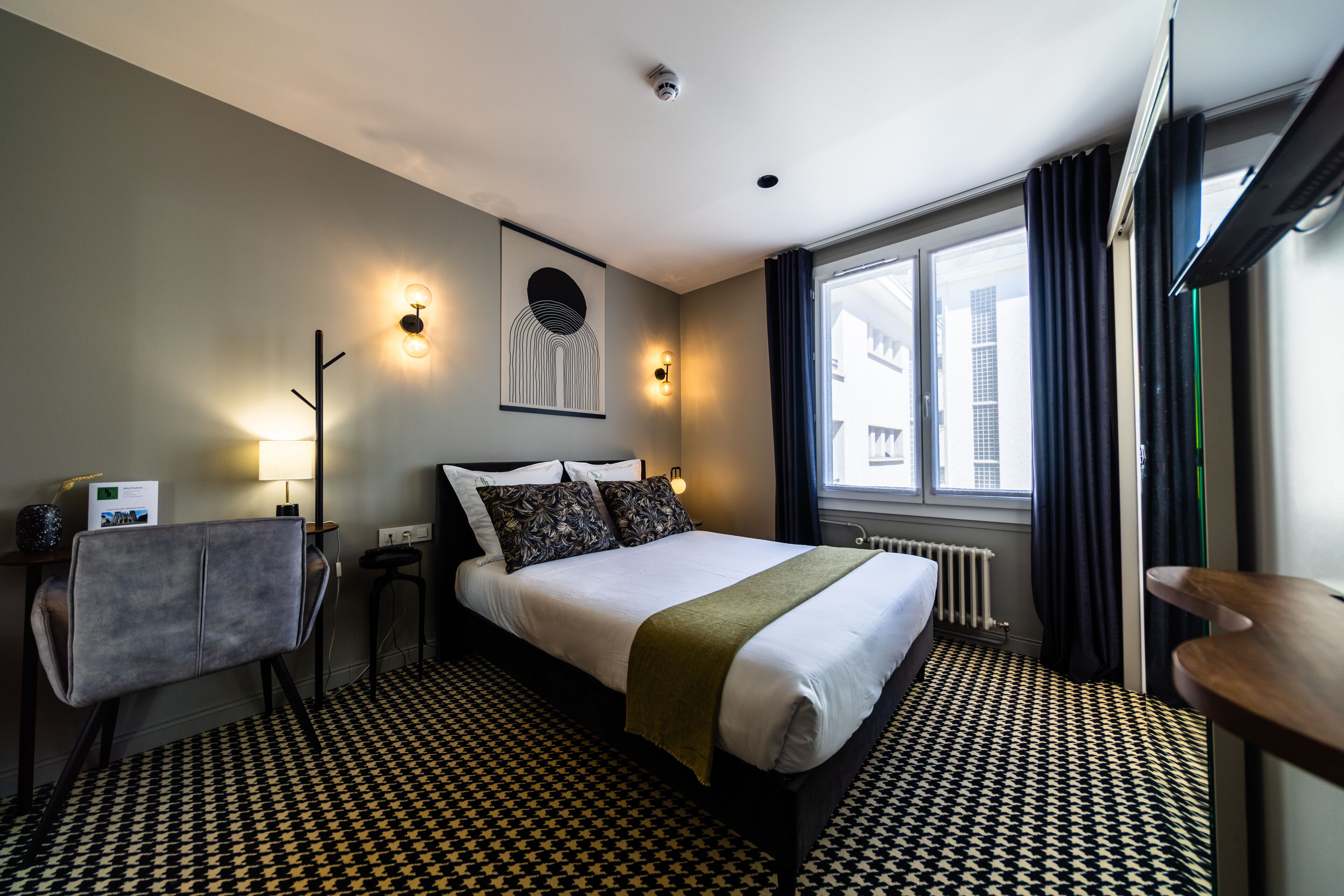 Habitación de hotel con una cama bien hecha, un escritorio con silla, una lámpara, una obra de arte en la pared y una ventana con cortinas.