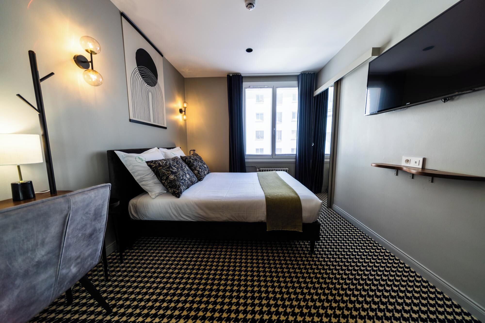 A hotel room with a bed, a TV mounted on the wall, a window with curtains, and a patterned floor.