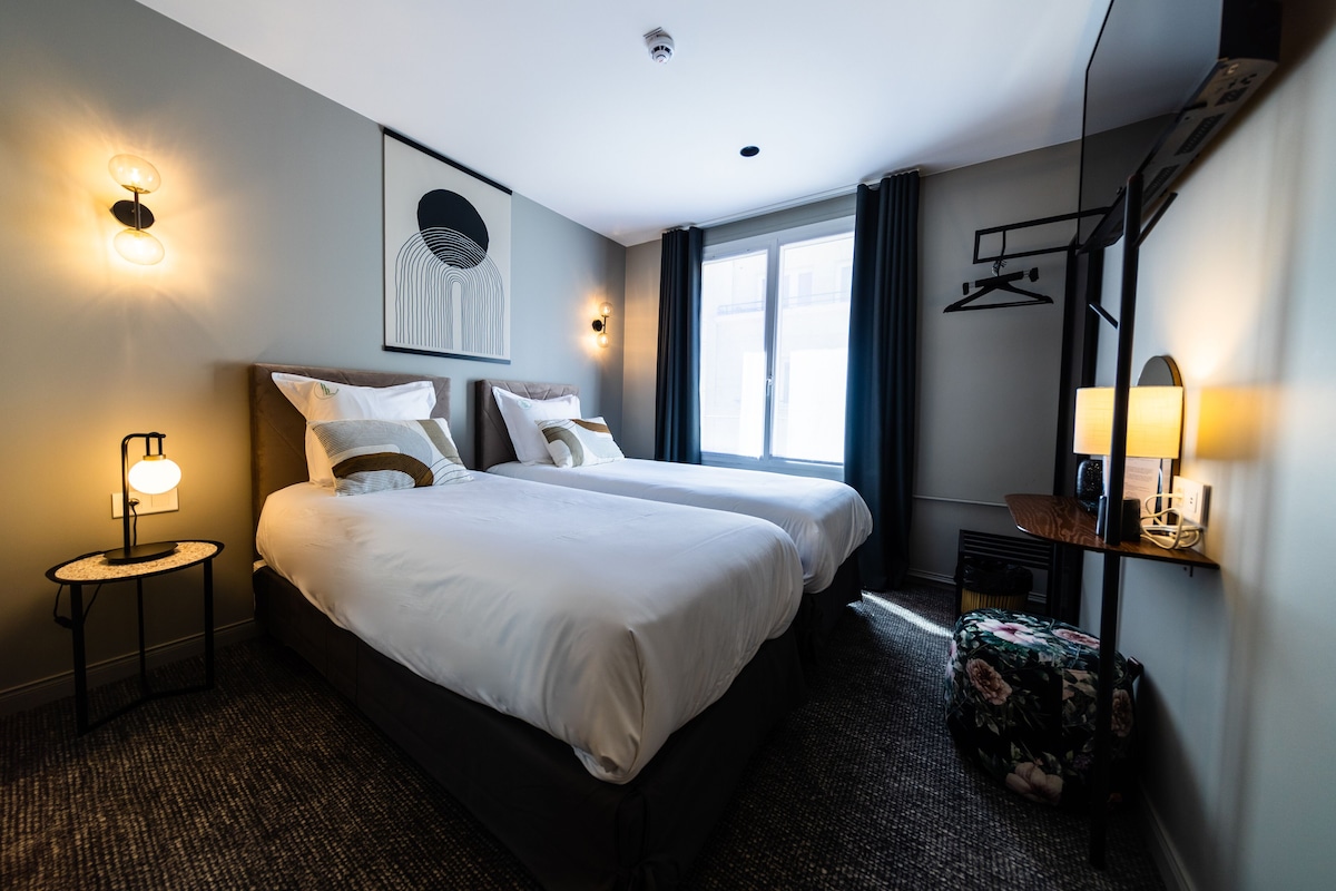 Une chambre d’hôtel avec deux lits, une petite table ronde, une lampe, un téléviseur fixé au mur et une fenêtre avec des rideaux.