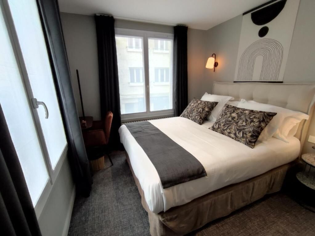 A hotel room with a large bed, a desk, a chair, a window with curtains, and a wall-mounted light fixture.