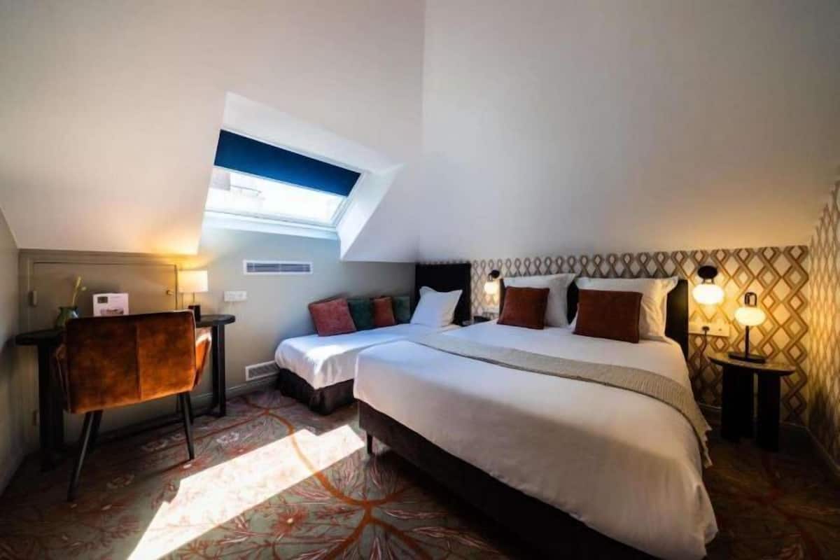 A hotel room with a large bed, two bedside tables, a desk with a chair, a small window, and patterned wallpaper.