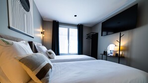 Habitación de hotel con dos camas, un televisor de pantalla plana montado en la pared, una mesita de noche con lámpara y una ventana con cortinas.