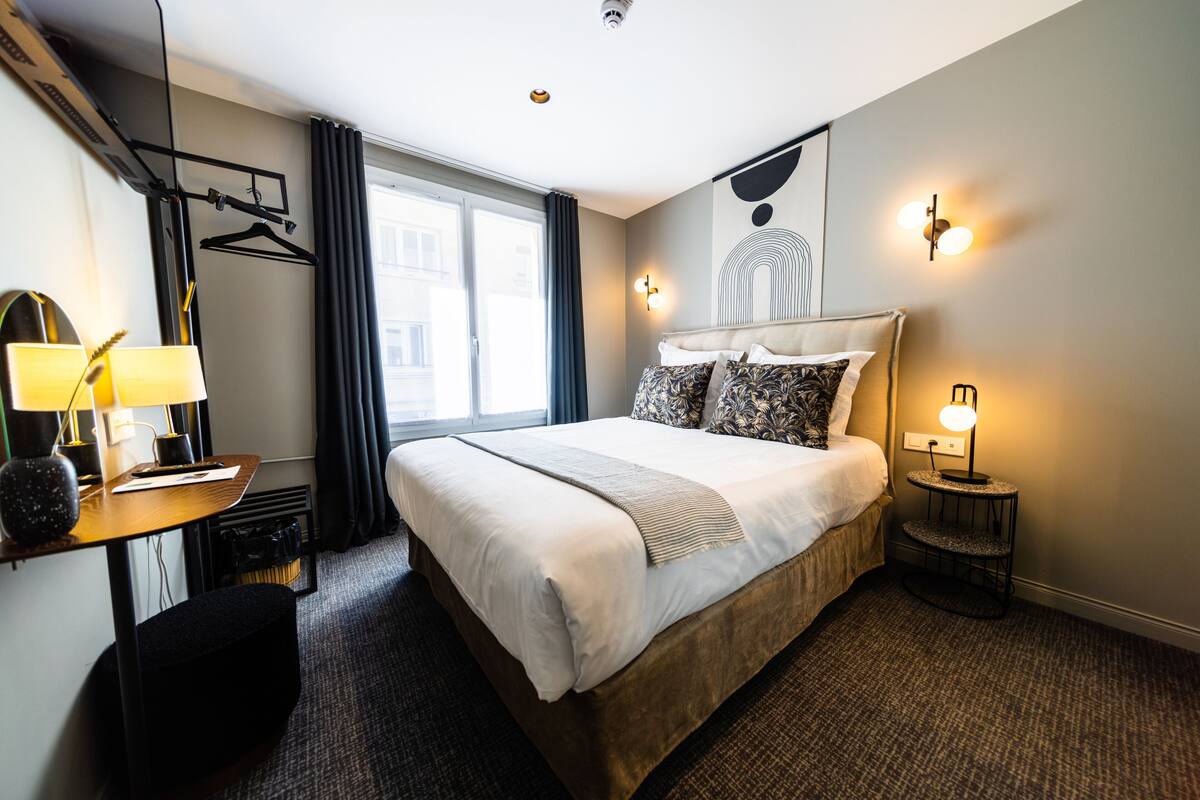 A hotel room with a large bed, a desk with a lamp, a chair, a coat rack, and a window with curtains.