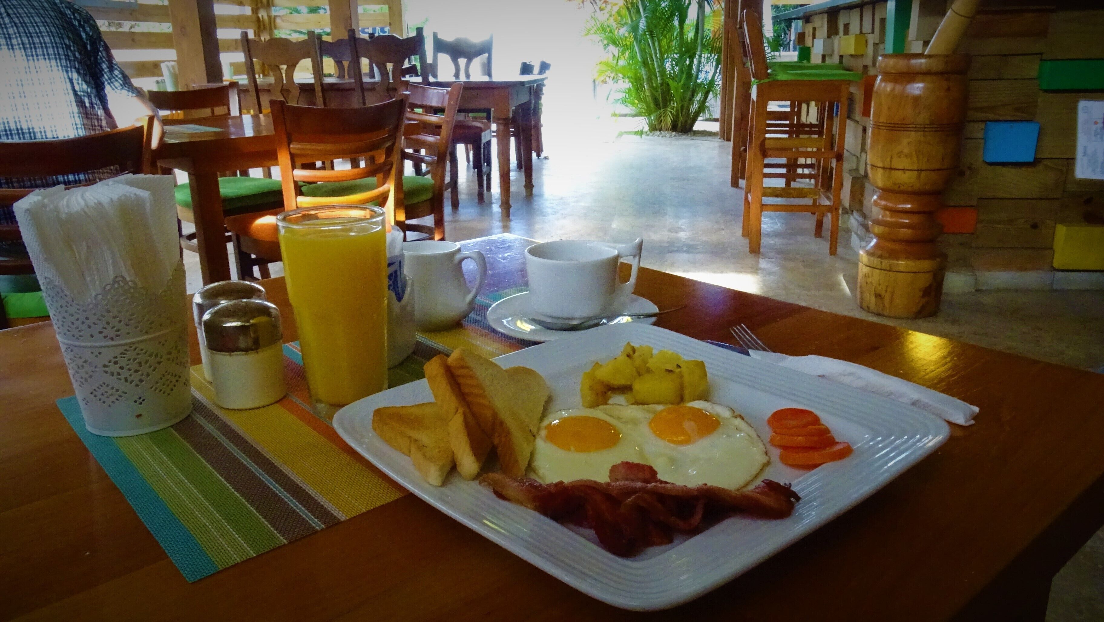 Una mesa de desayuno con huevos, tocino, tostadas y fruta. Hay tazas y una jarra de jugo de naranja.