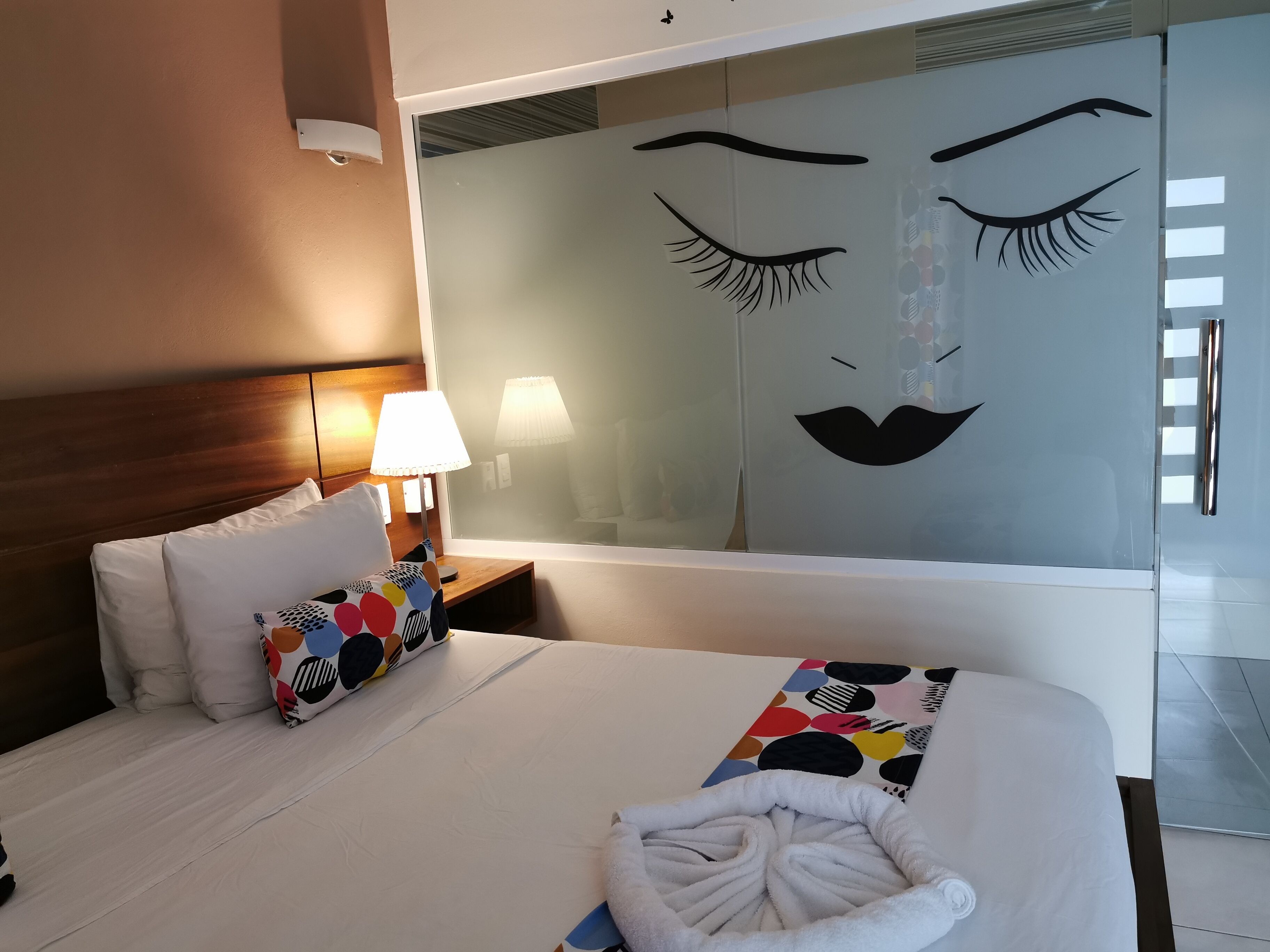 Ein Hotelzimmer mit Bett, Kissen, einer Nachttischlampe, einem großen Spiegel und einem Handtuch auf dem Bett.