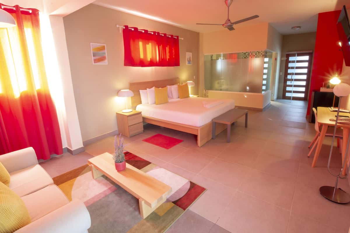 Ein modernes Hotelzimmer mit einem großen Bett, Nachttischen mit Lampen, einer Sitzecke und einem Essbereich mit Tisch und Stühlen sowie einem großen Spiegel.