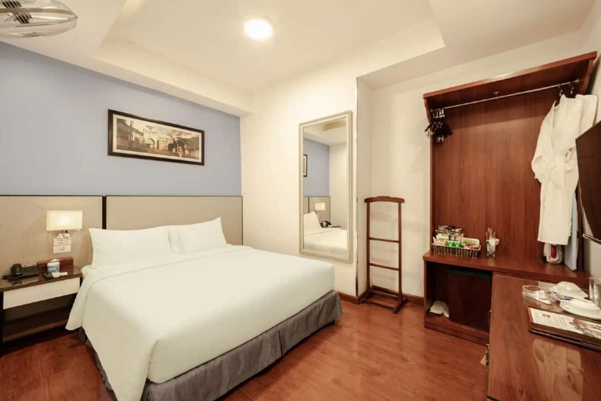 A hotel room with a large bed, a wooden wardrobe, a bedside table with a lamp, and a framed picture on the wall.
