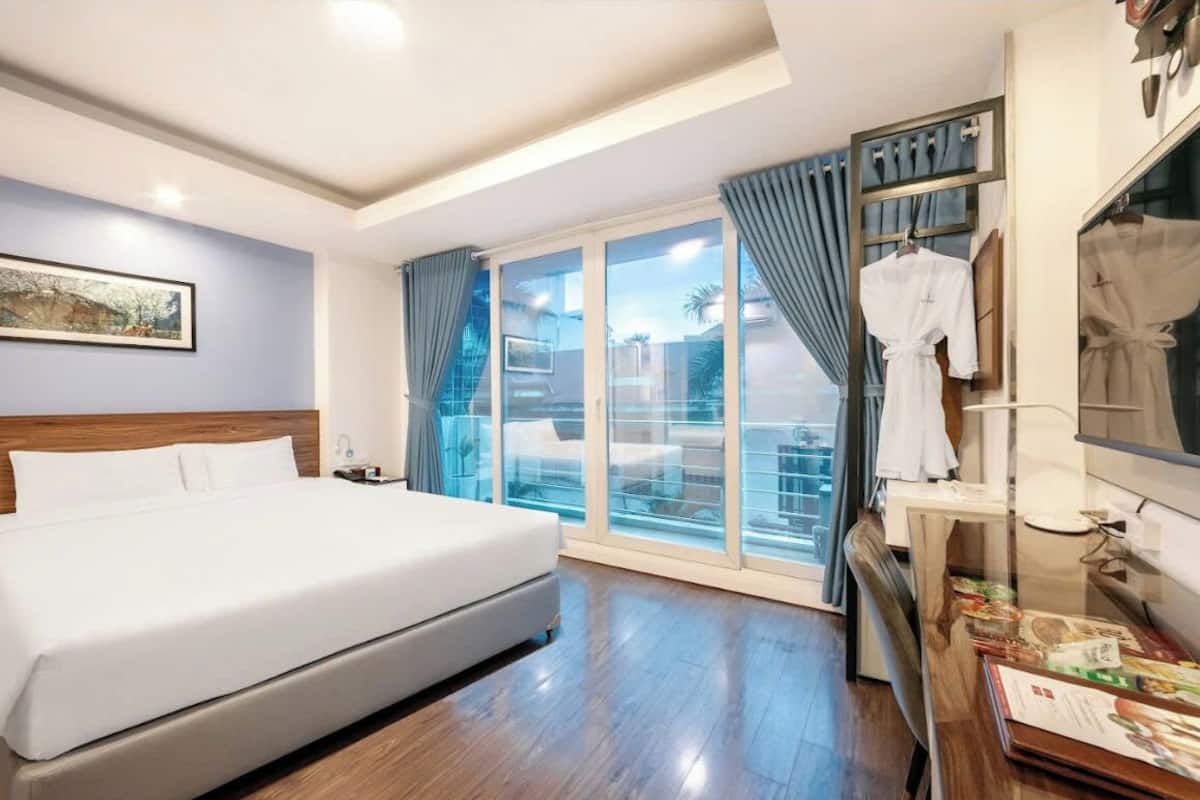 A hotel room with a large bed, a desk with a lamp, a wardrobe, and a sliding glass door to the balcony.