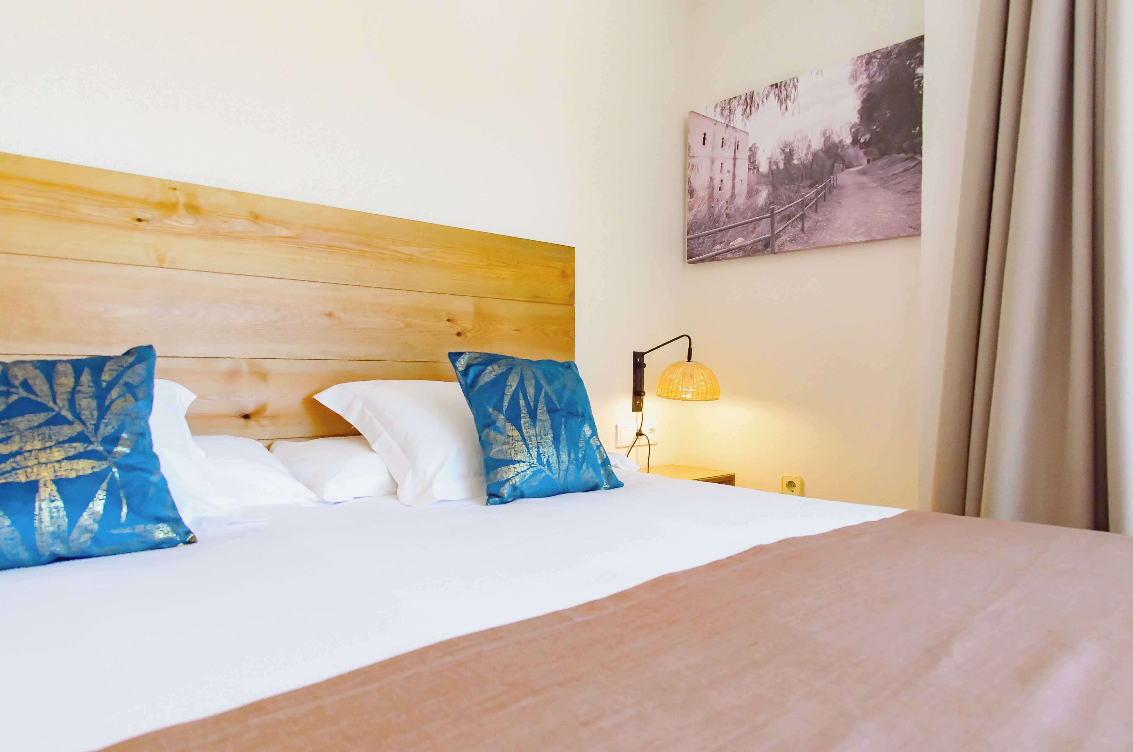 A neatly made bed with white and blue pillows, a wooden headboard, a wall-mounted lamp, and a framed picture.