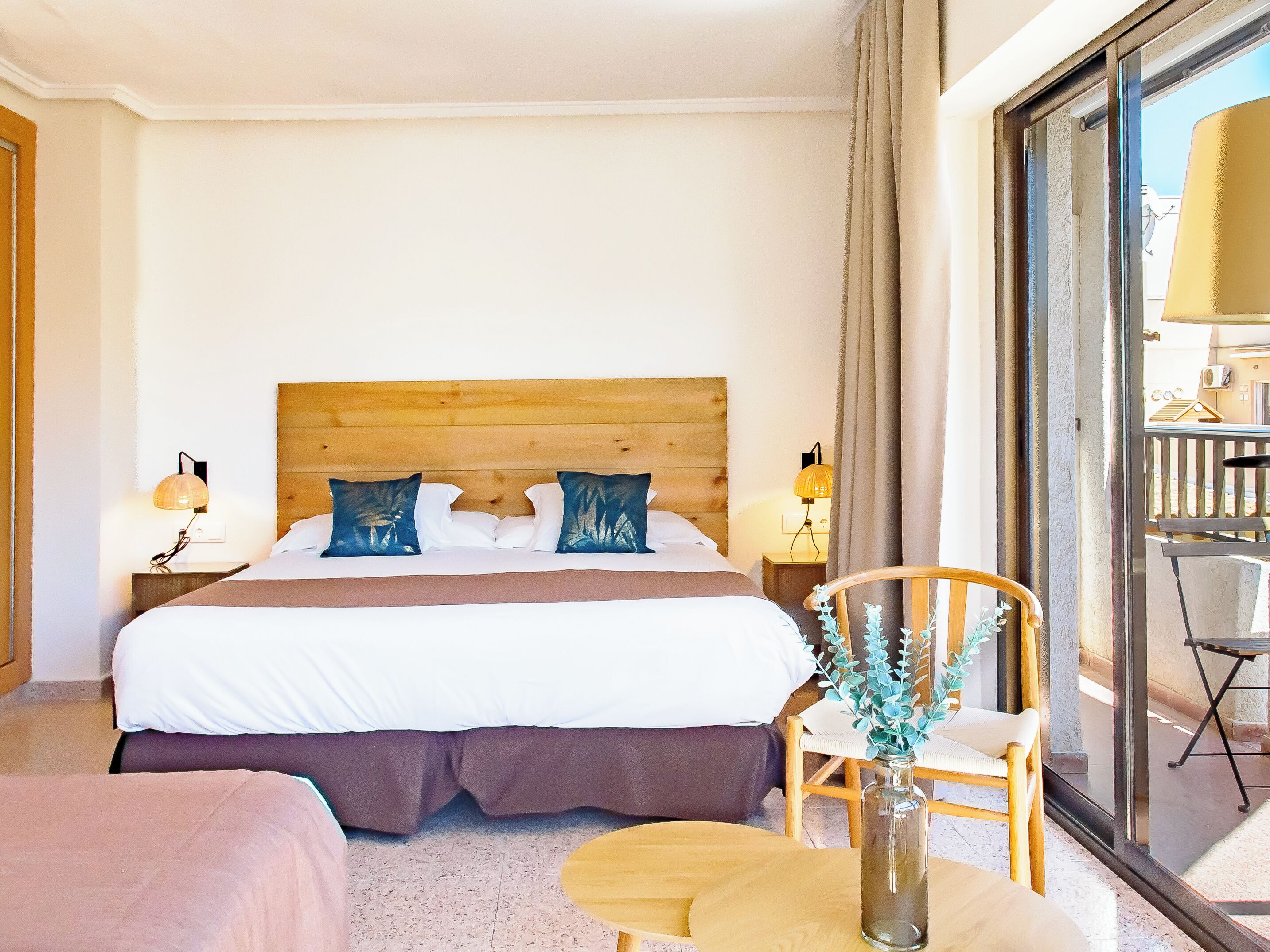 Habitación de hotel con una cama grande, dos mesitas de noche con lámparas, una silla, una pequeña mesa y vistas al balcón exterior.