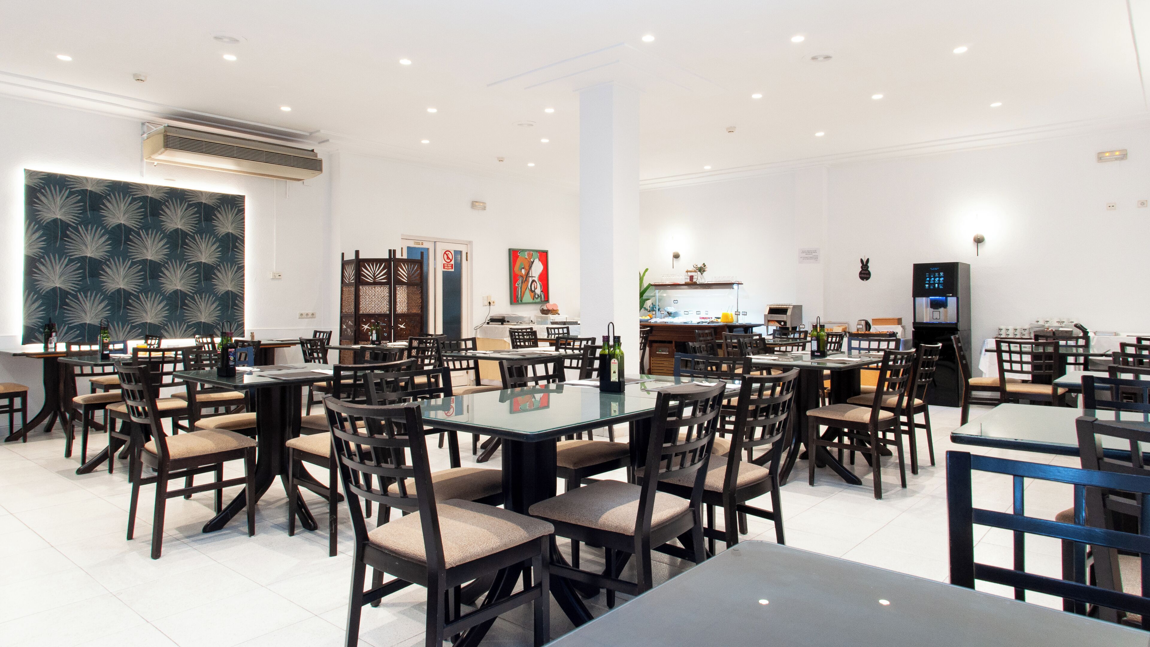 Uma sala de jantar espaçosa com várias mesas e cadeiras, cozinha ao fundo e uma parede decorativa com padrão de folhas.