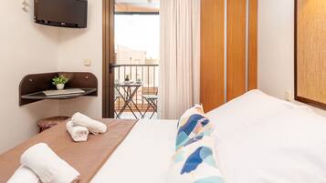 A hotel room with a bed, a desk with a laptop, a small table with a plant, and a balcony with a table and chairs.