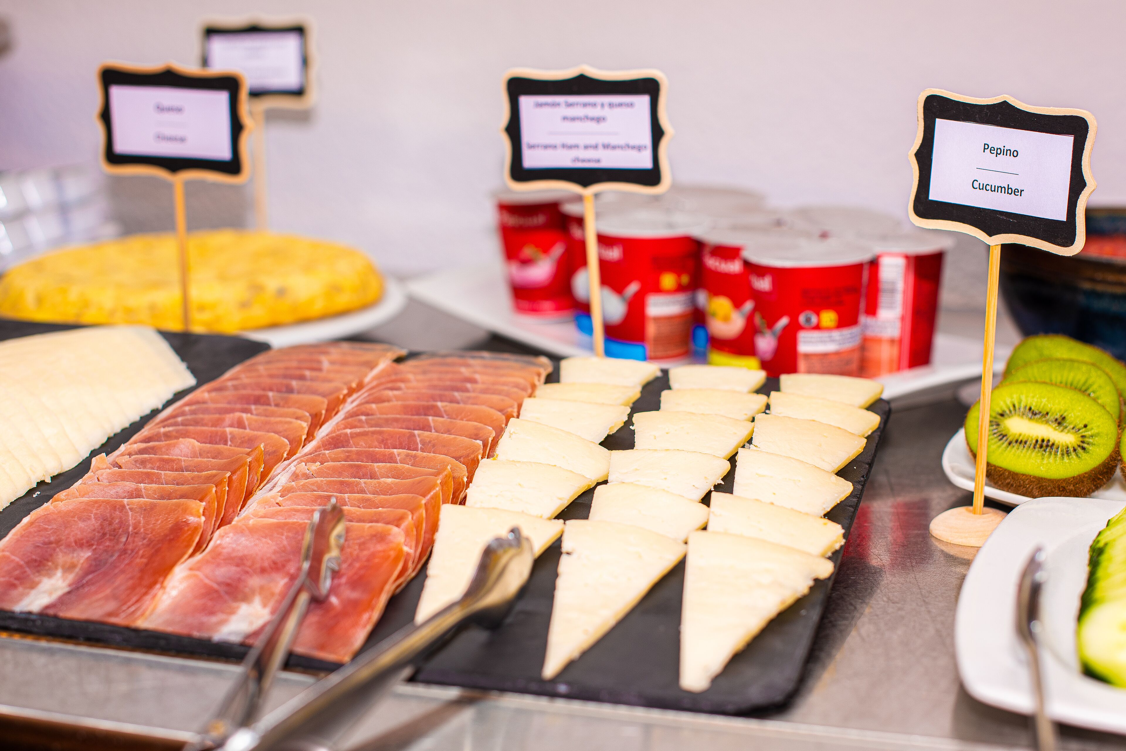 Buffet com diversos queijos, embutidos e frutas como kiwi e pepino.
