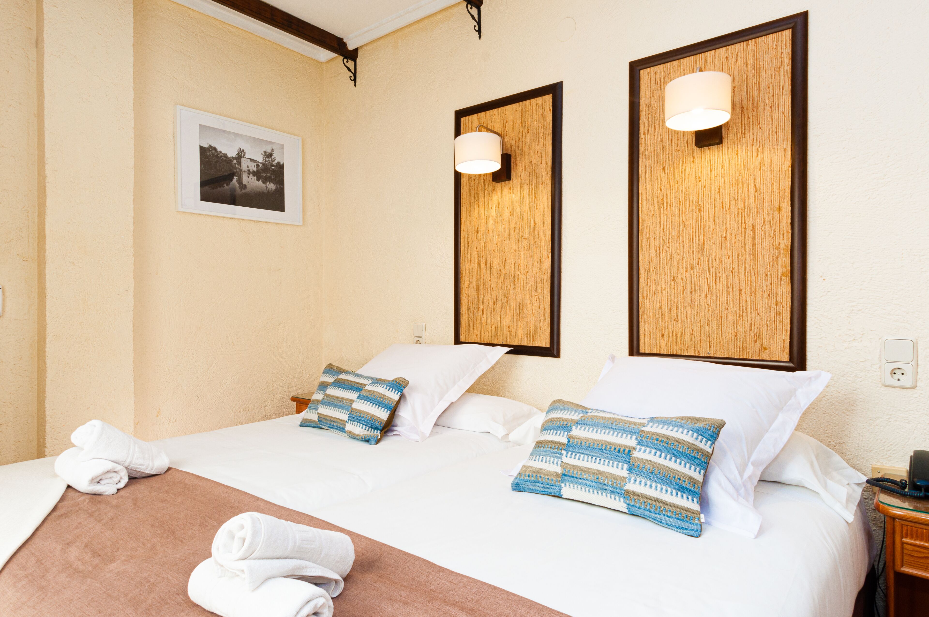 Quarto de hotel com duas camas, cada uma com roupa de cama branca e almofadas estampadas. Há duas luminárias fixadas na parede e um quadro emoldurado.