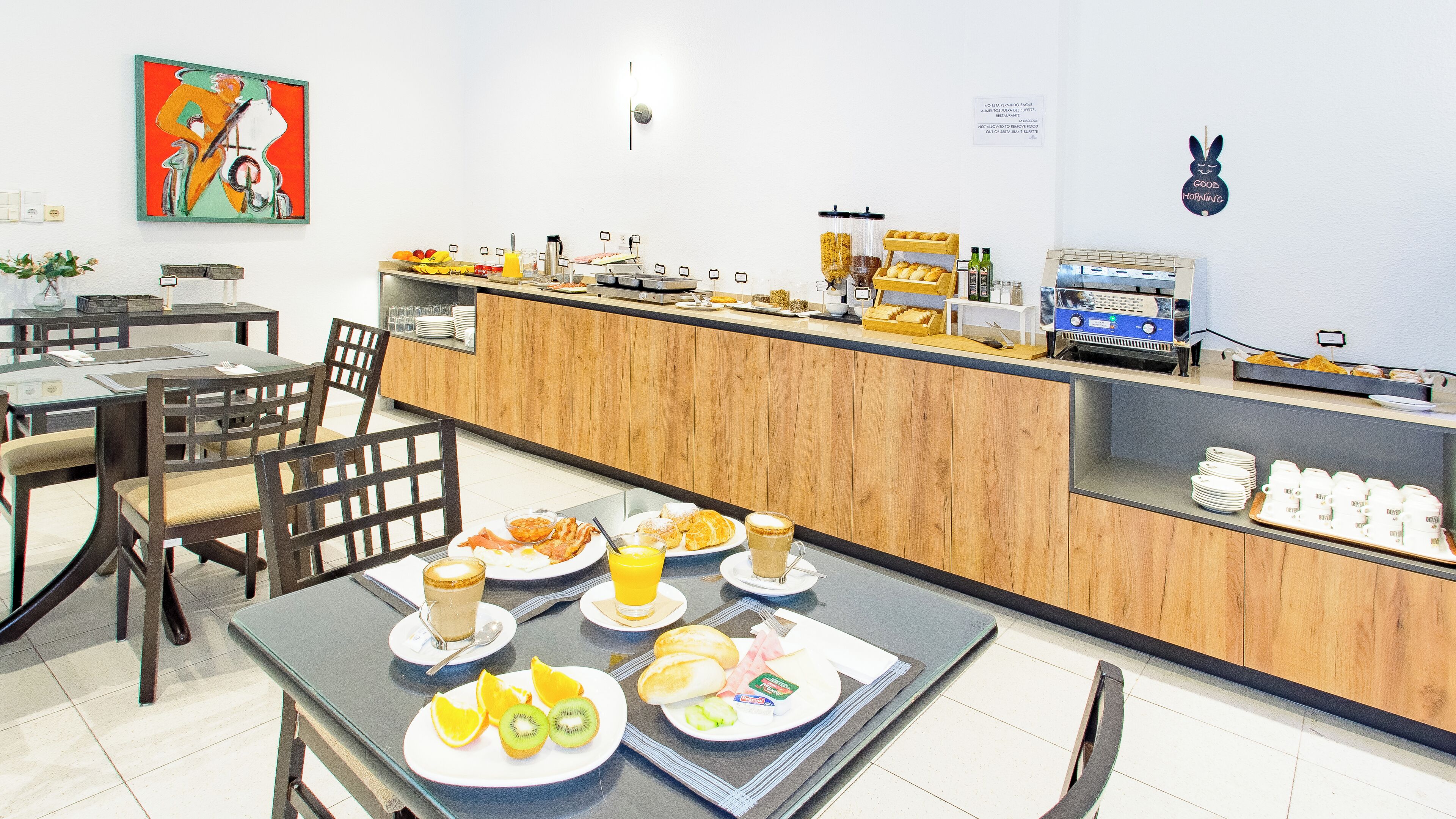 A breakfast buffet with various food items and beverages, a coffee machine, and a wall-mounted sign.