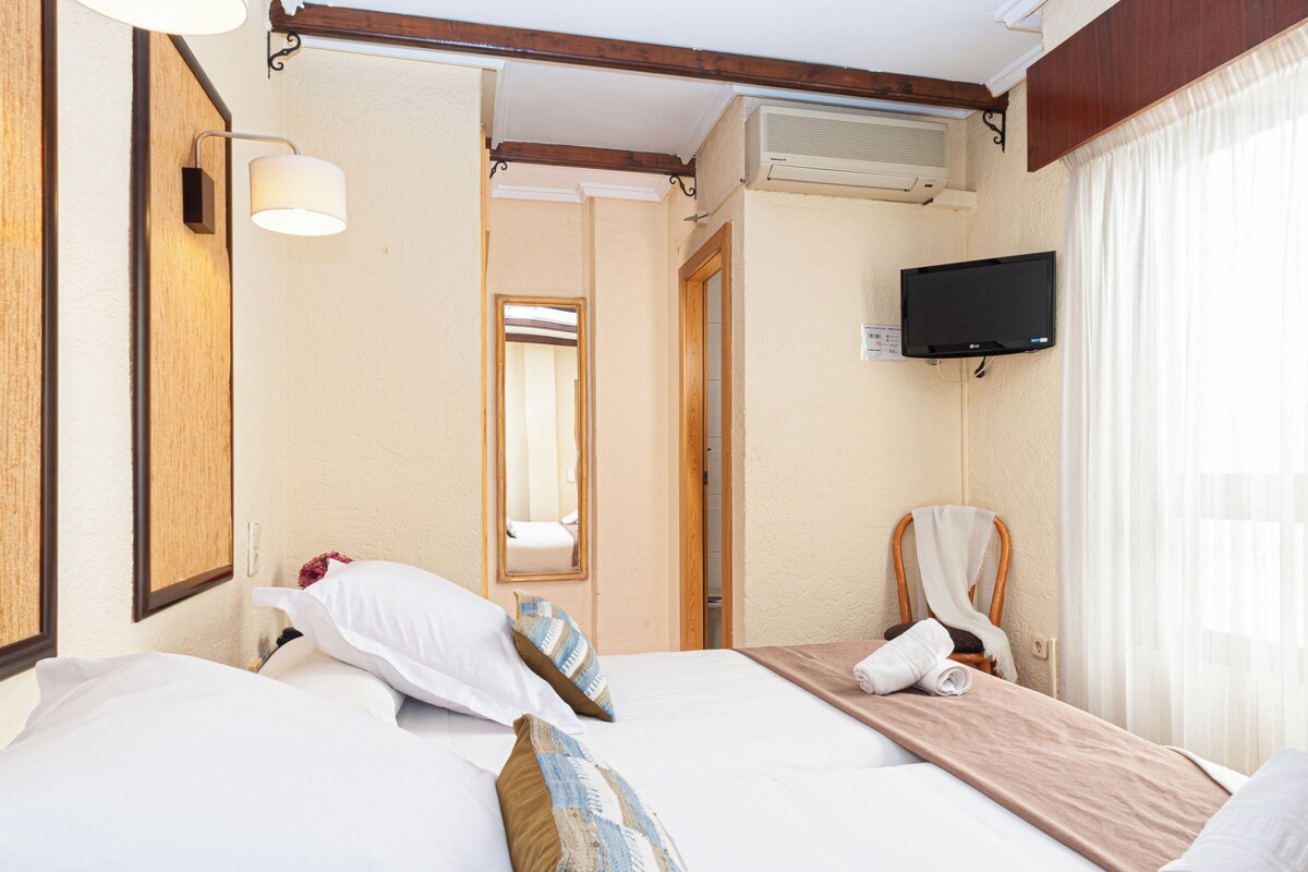 A hotel room with a bed, a TV mounted on the wall, a mirror, and a window with curtains.