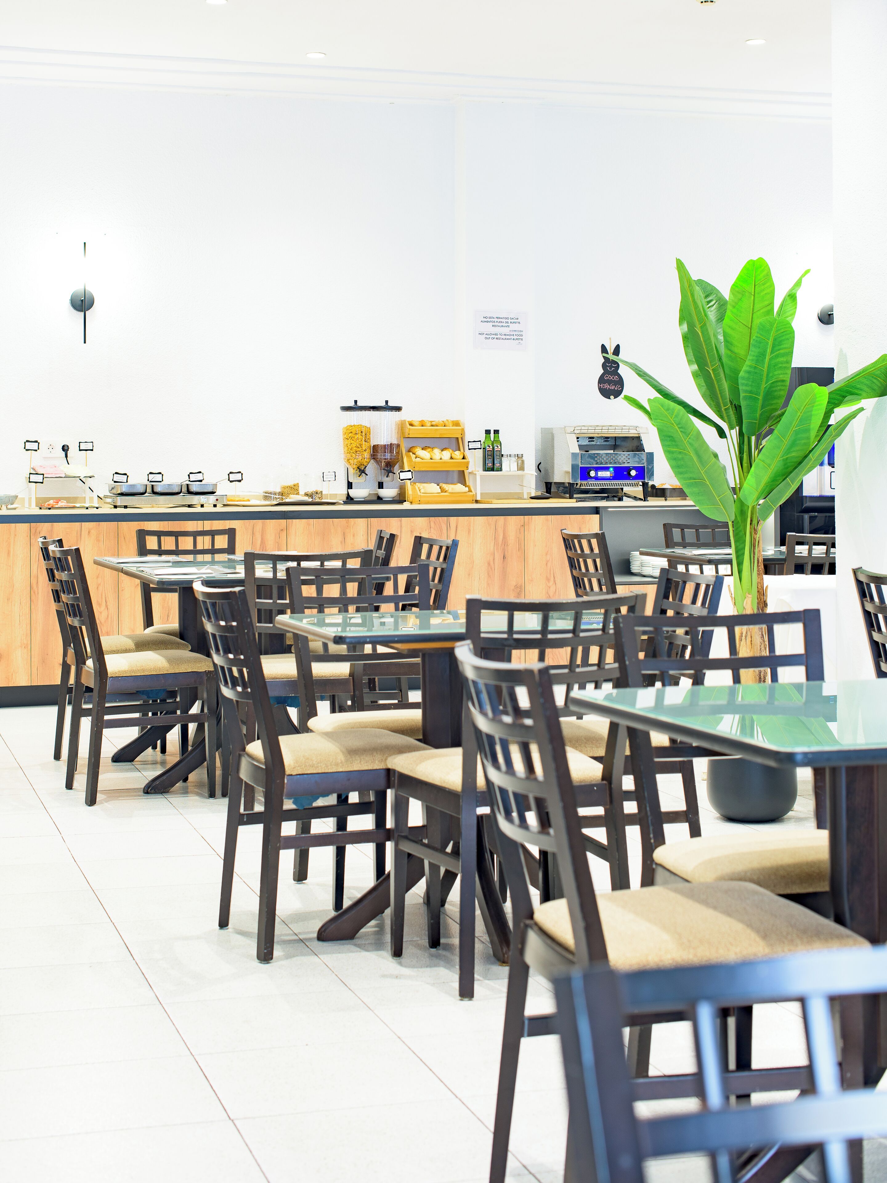 Un restaurant amb taules i cadires de fusta, una barra amb maquinària de cuina i una planta verda gran.