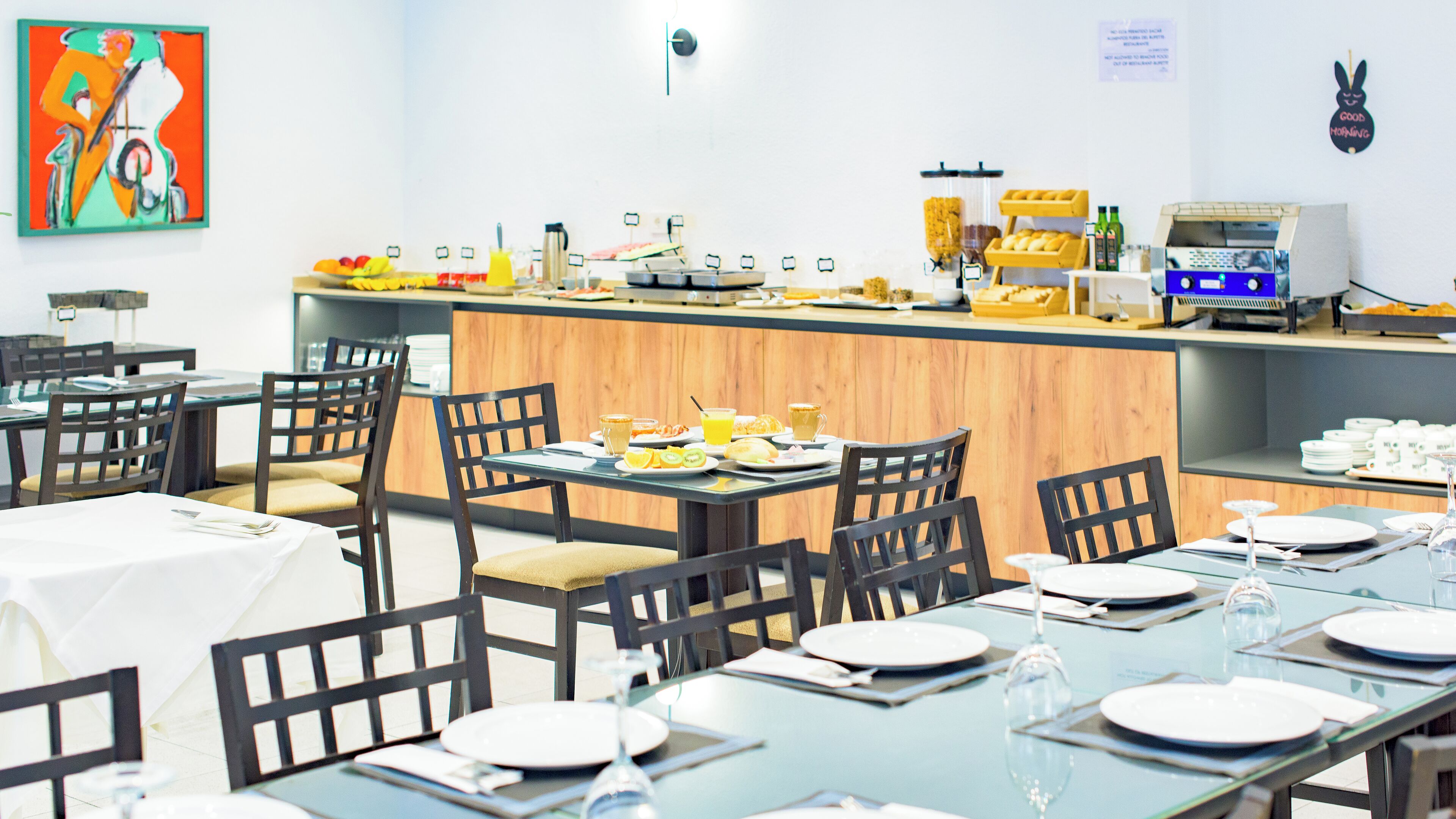 A modern dining area with glass tables and chairs, a buffet counter with various food items, and a wall with a colorful painting.