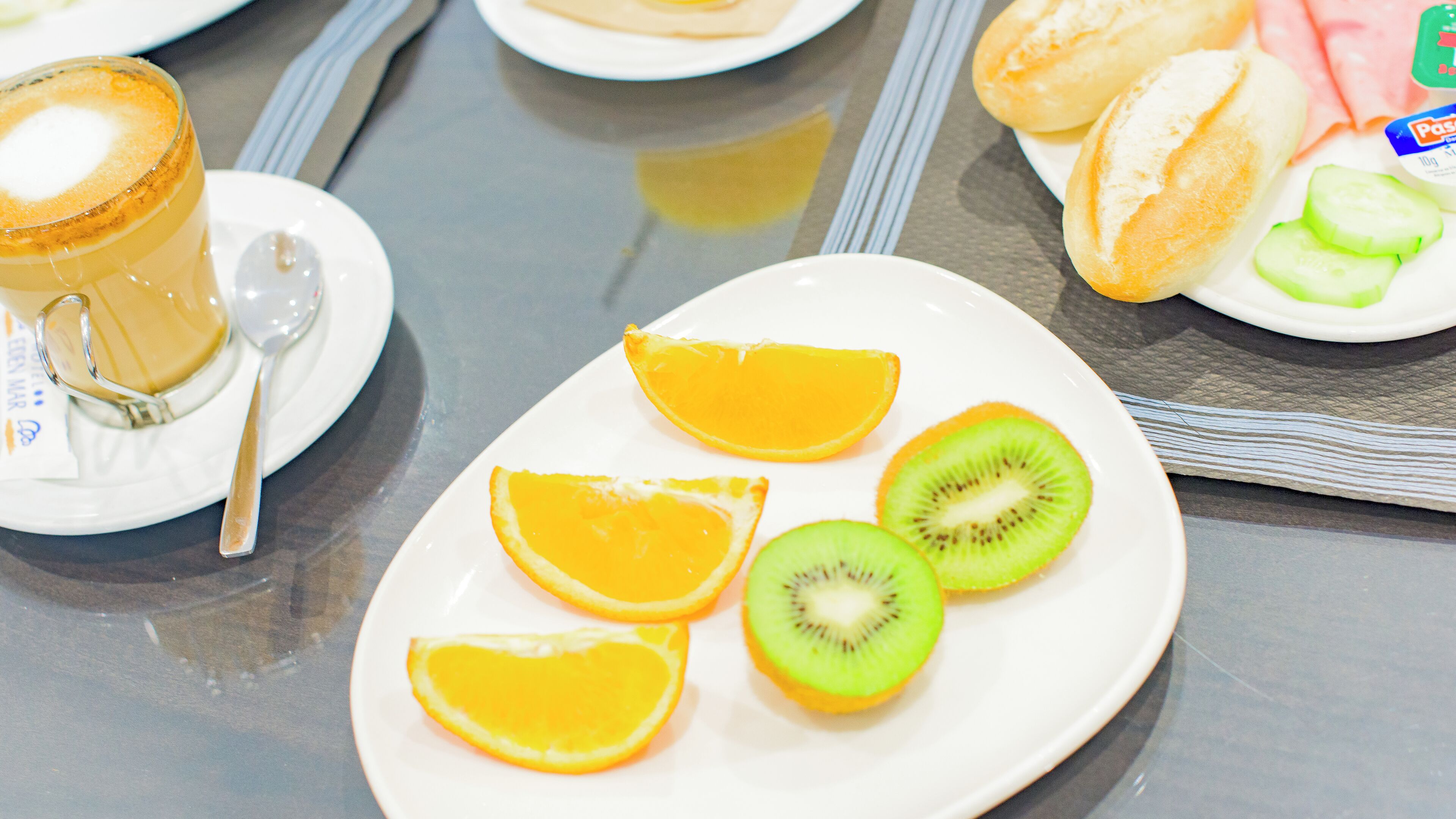 A breakfast setting with a plate of sliced oranges and kiwi, a glass of coffee, and a plate of bread and cheese.
