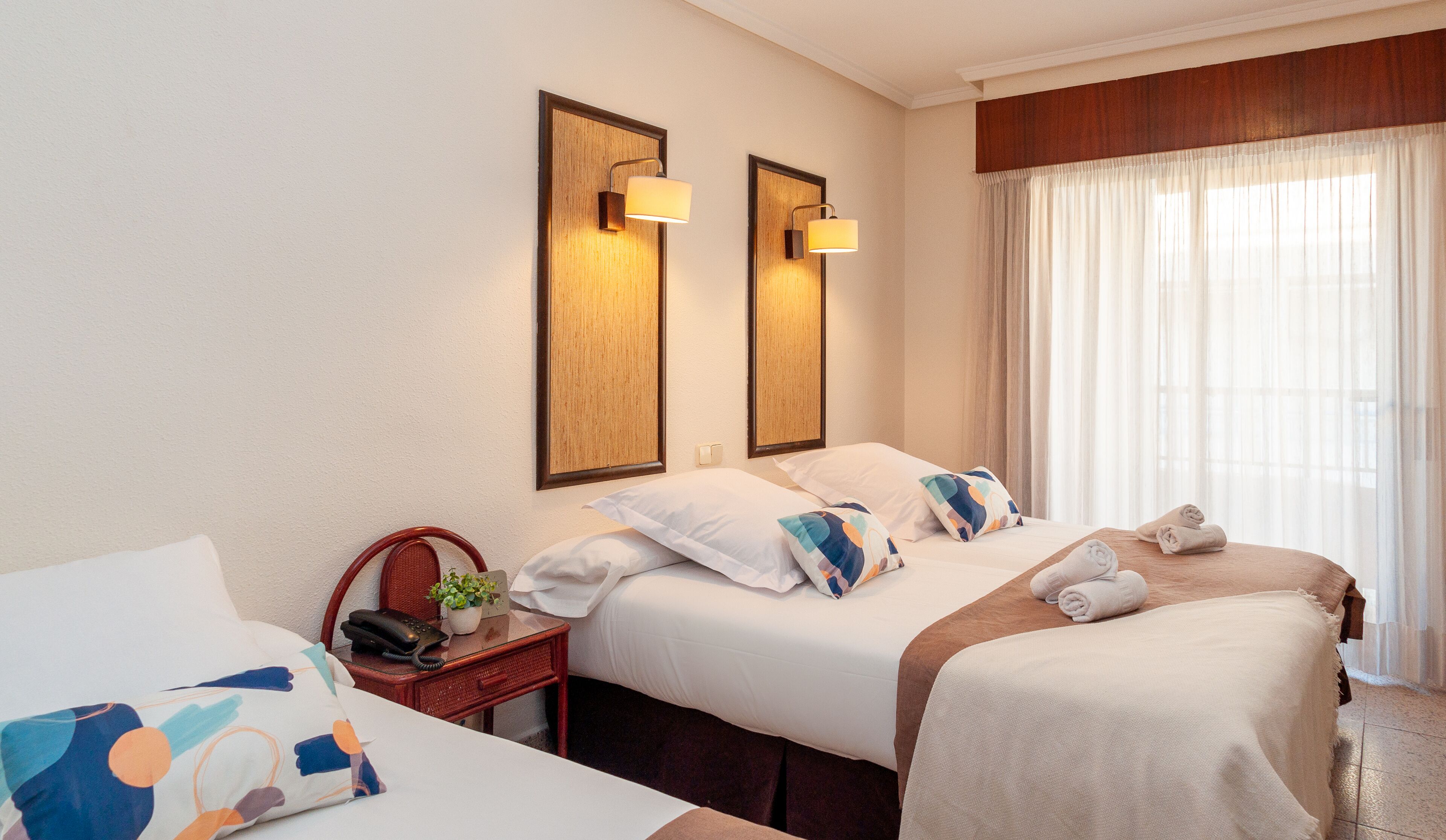 Une chambre d’hôtel avec deux lits, une table de chevet avec un téléphone et une plante, et une fenêtre avec des rideaux.
