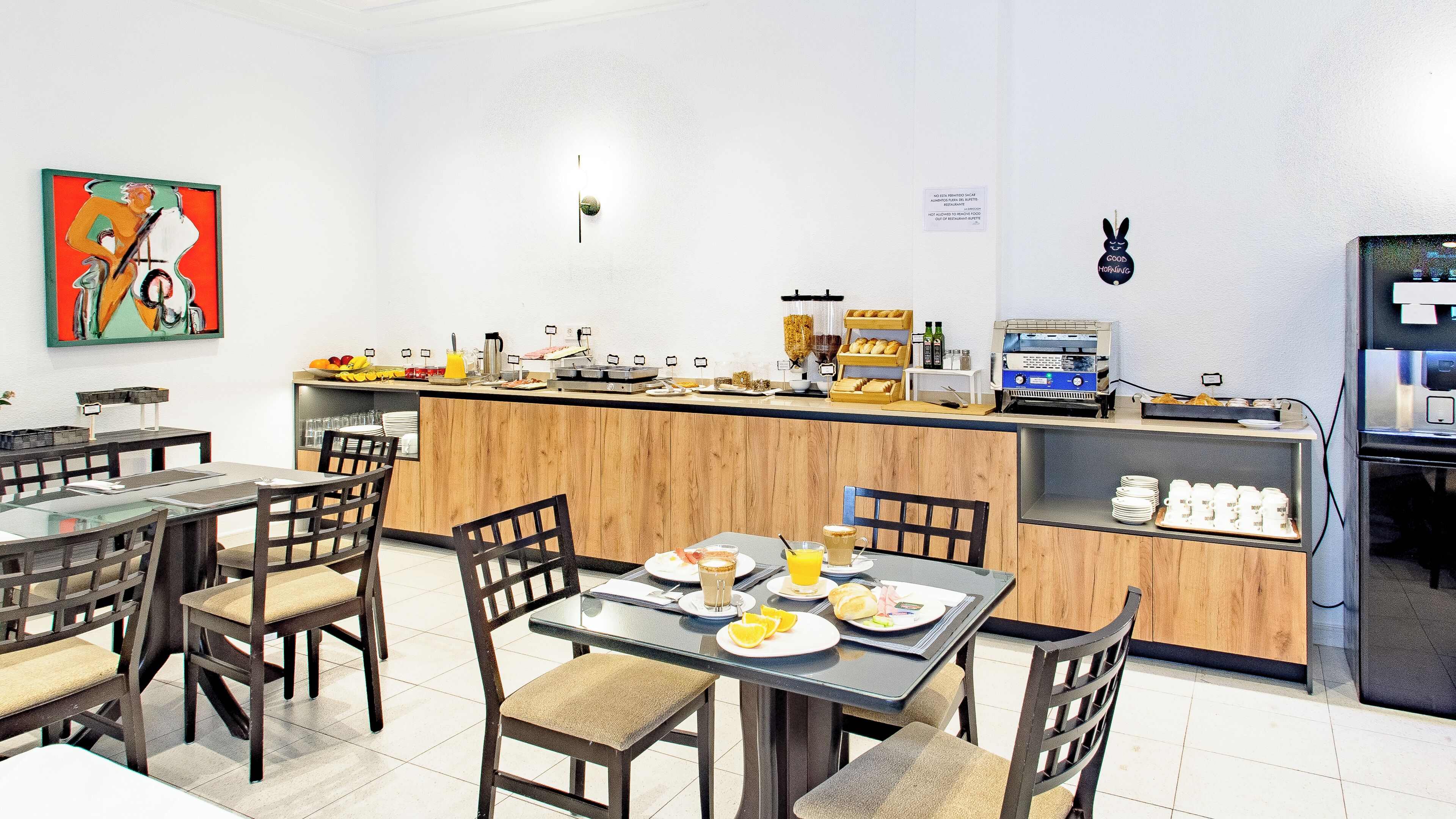 Un espace moderne dédié au petit-déjeuner, avec des tables et des chaises en bois, un comptoir proposant divers articles pour le petit-déjeuner et une machine à café.