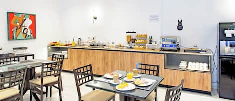 Un espace moderne dédié au petit-déjeuner, avec des tables et des chaises en bois, un comptoir proposant divers articles pour le petit-déjeuner et une machine à café.