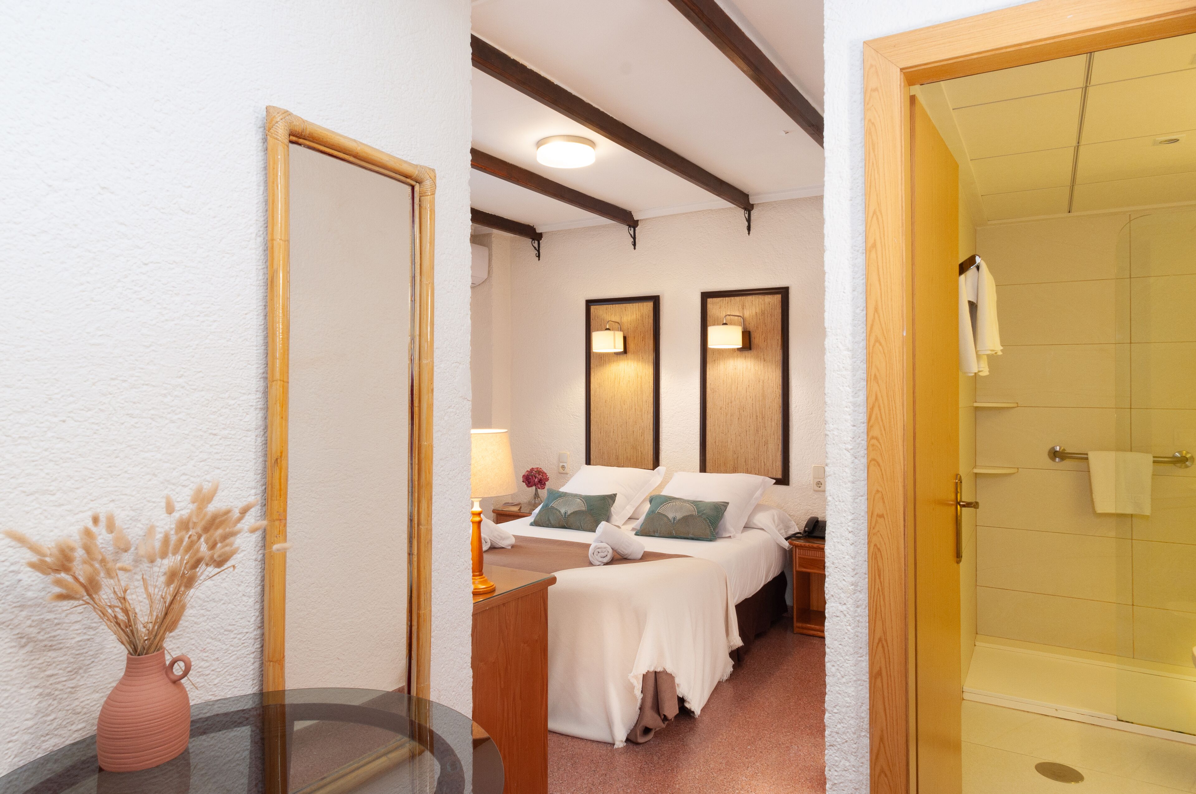 A hotel room with a bed, bedside lamps, a nightstand, a vase with dried flowers, and a glass shower enclosure.