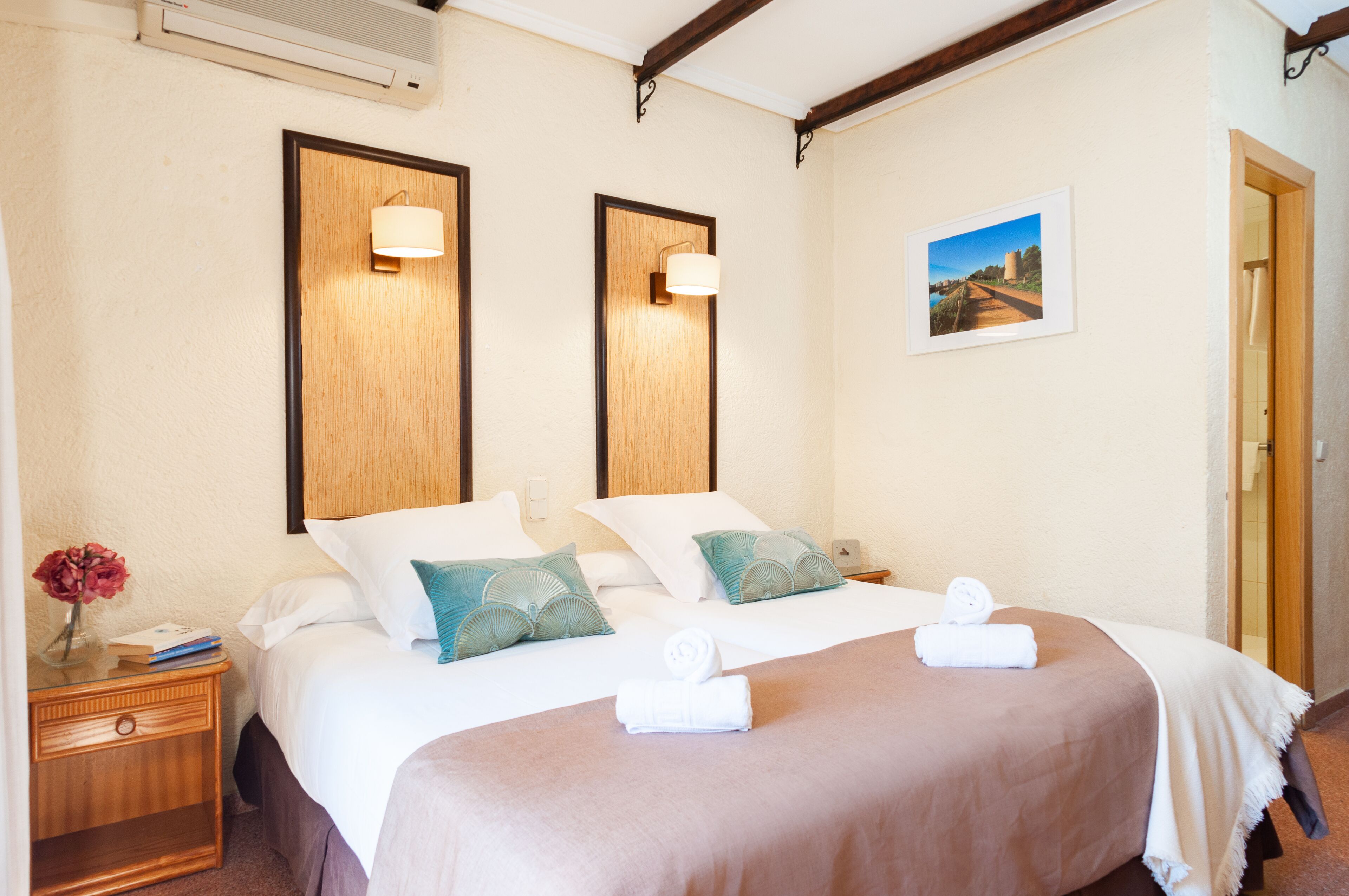 Une chambre d’hôtel avec deux lits, chacun doté d’une tête de lit en bois, de draps blancs et d’oreillers turquoise. On y trouve des lampes fixées au mur, un tableau encadré et une table de chevet avec un vase de fleurs et un livre.