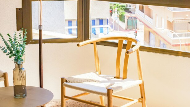 A room with a wooden chair, a small table with a vase, and a lamp. A large window offers a view of a beach and buildings.