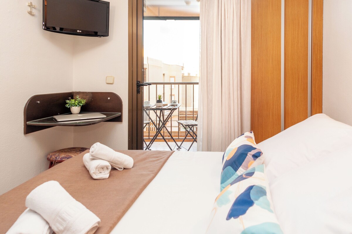 A hotel room with a bed, a desk with a laptop, a small table with a plant, and a balcony with a table and chairs.