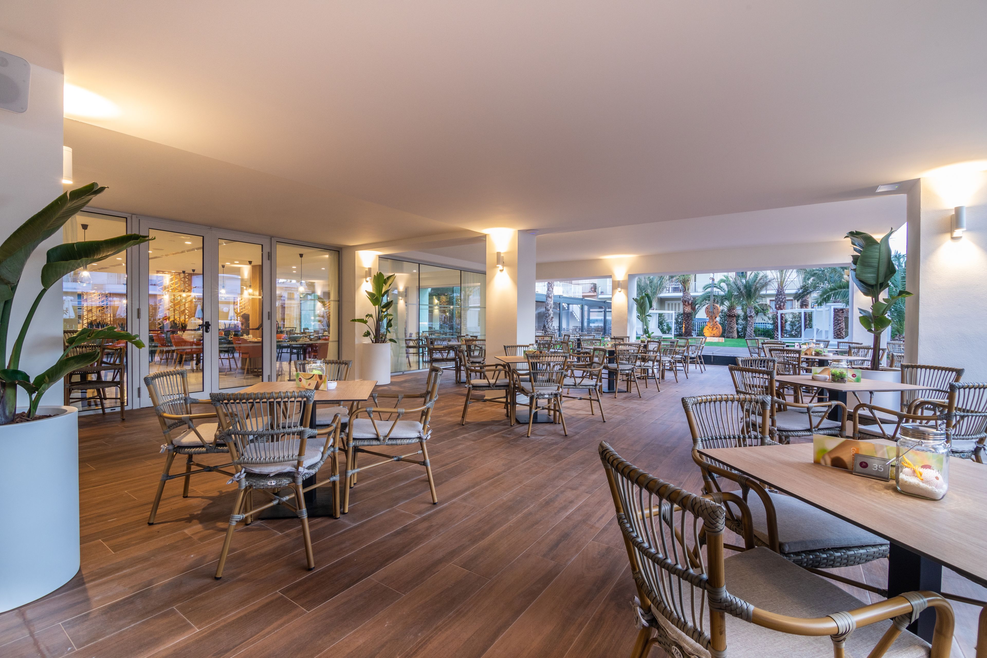 A spacious indoor dining area with wooden flooring and wicker chairs. Tables are arranged in rows, and there are large windows providing natural light.