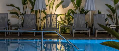 Zona de piscina con sillones de descanso y sombrillas, rodeada de plantas verdes grandes.