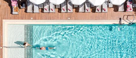 An aerial view of a swimming pool with a person swimming, lounge chairs, and umbrellas.