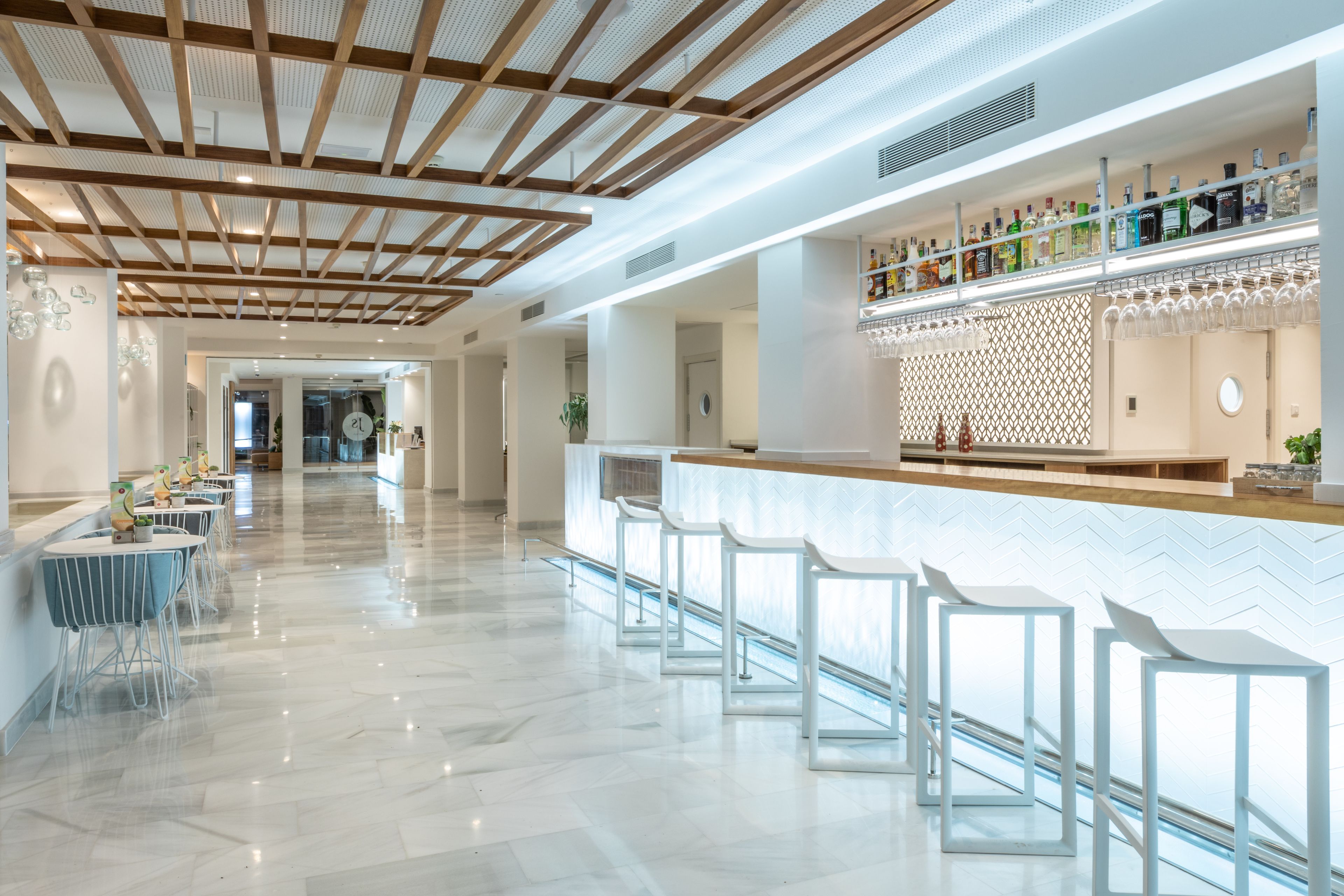 Un área de bar moderna con piso de mármol blanco, vigas de madera en el techo y una barra con taburetes.