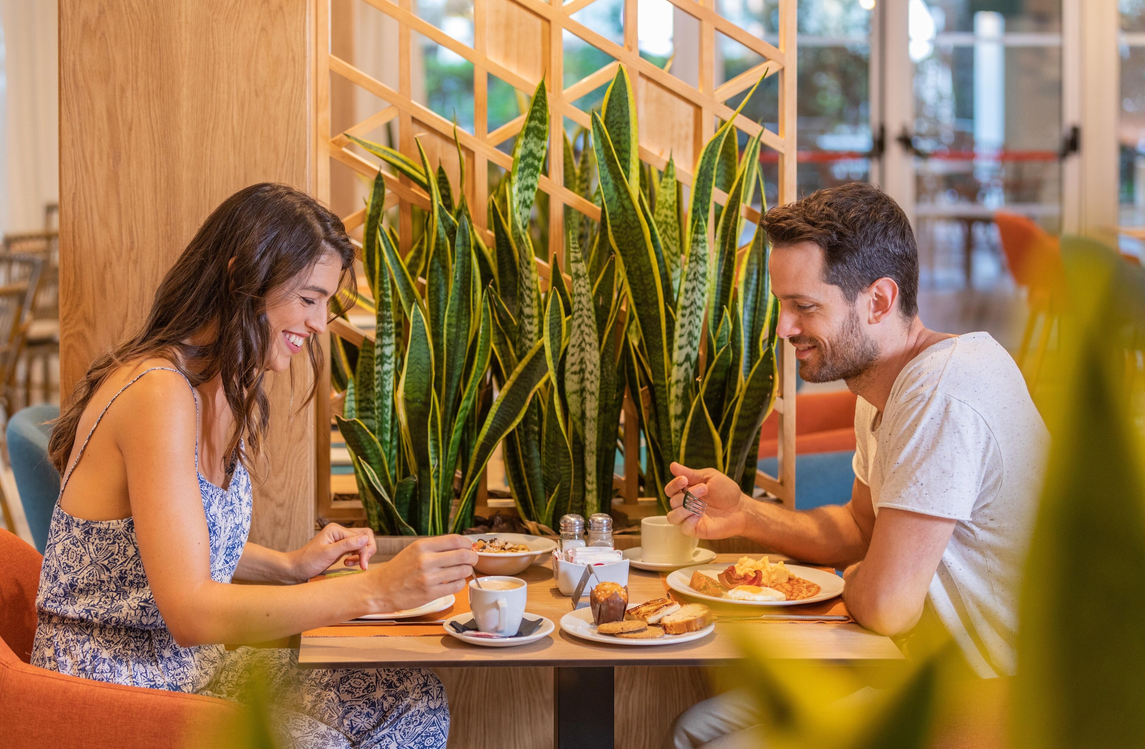 Duas pessoas jantando à mesa, com um divisória de madeira e plantas verdes ao fundo.