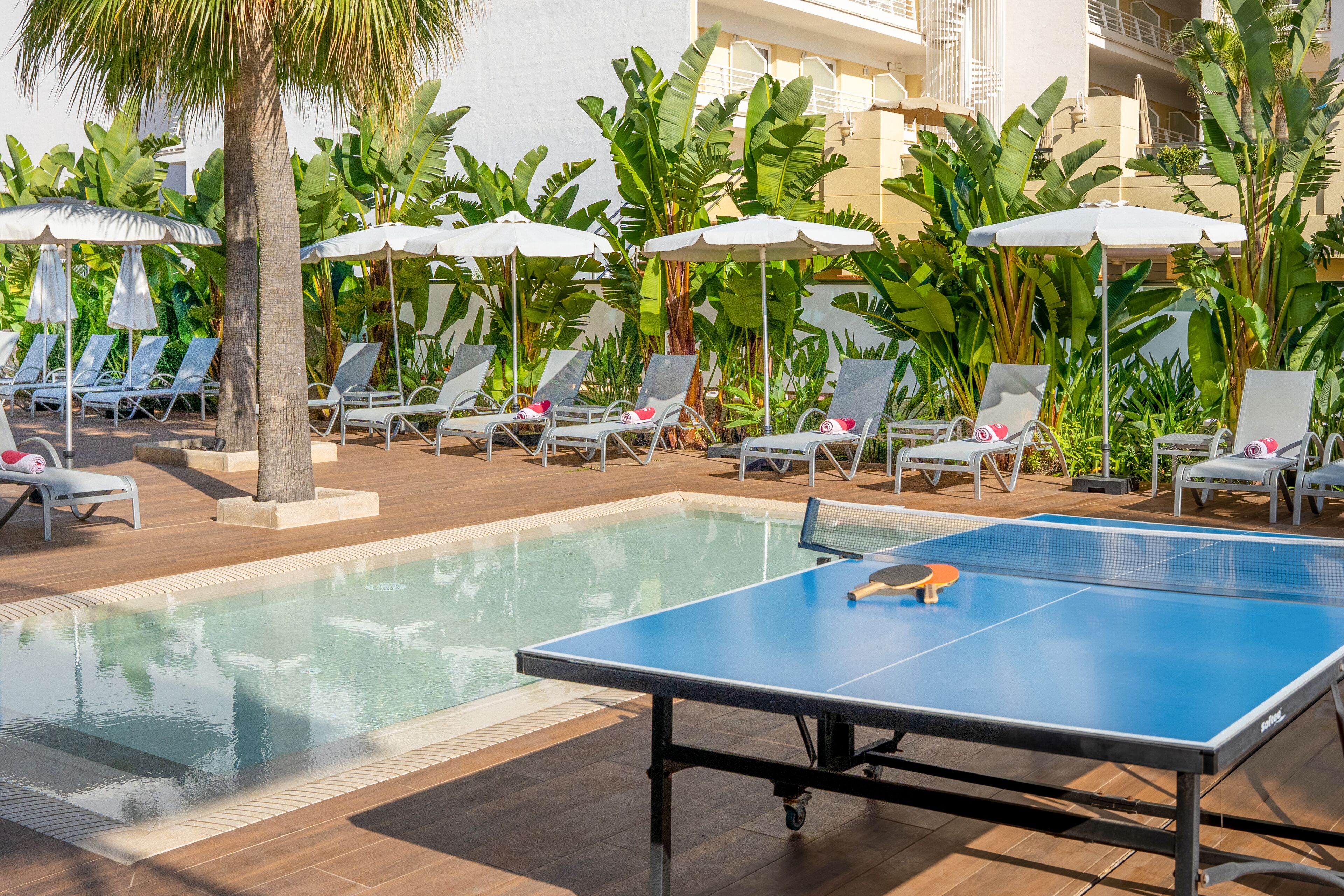 Un área de piscina con sillones de descanso y sombrillas, una mesa de ping pong y exuberante vegetación tropical.