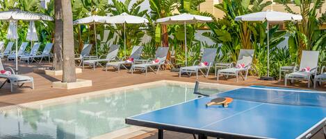 Un área de piscina con sillones de descanso y sombrillas, una mesa de ping pong y exuberante vegetación tropical.