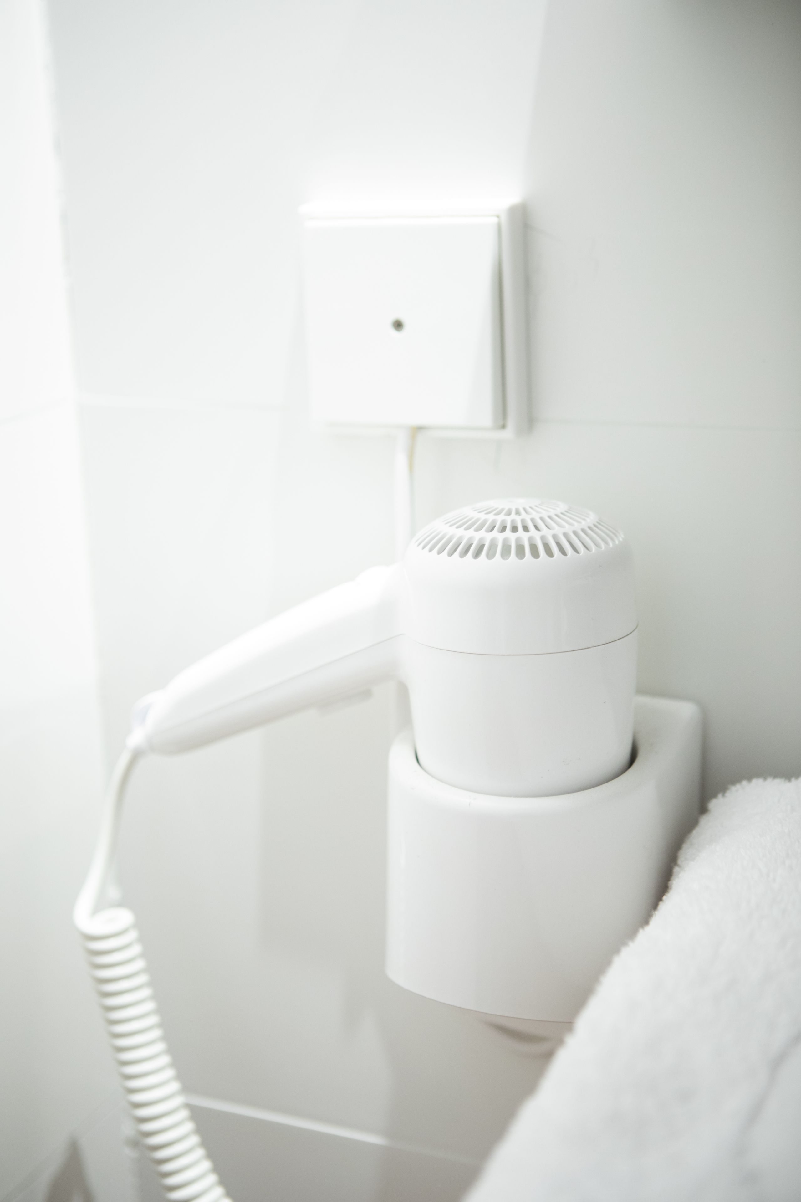 A white hair dryer mounted on a wall with a towel hanging nearby.
