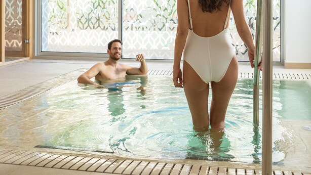Una piscina interior con agua cristalina, suelo de baldosas y vista a un edificio con cerca decorativa exterior.