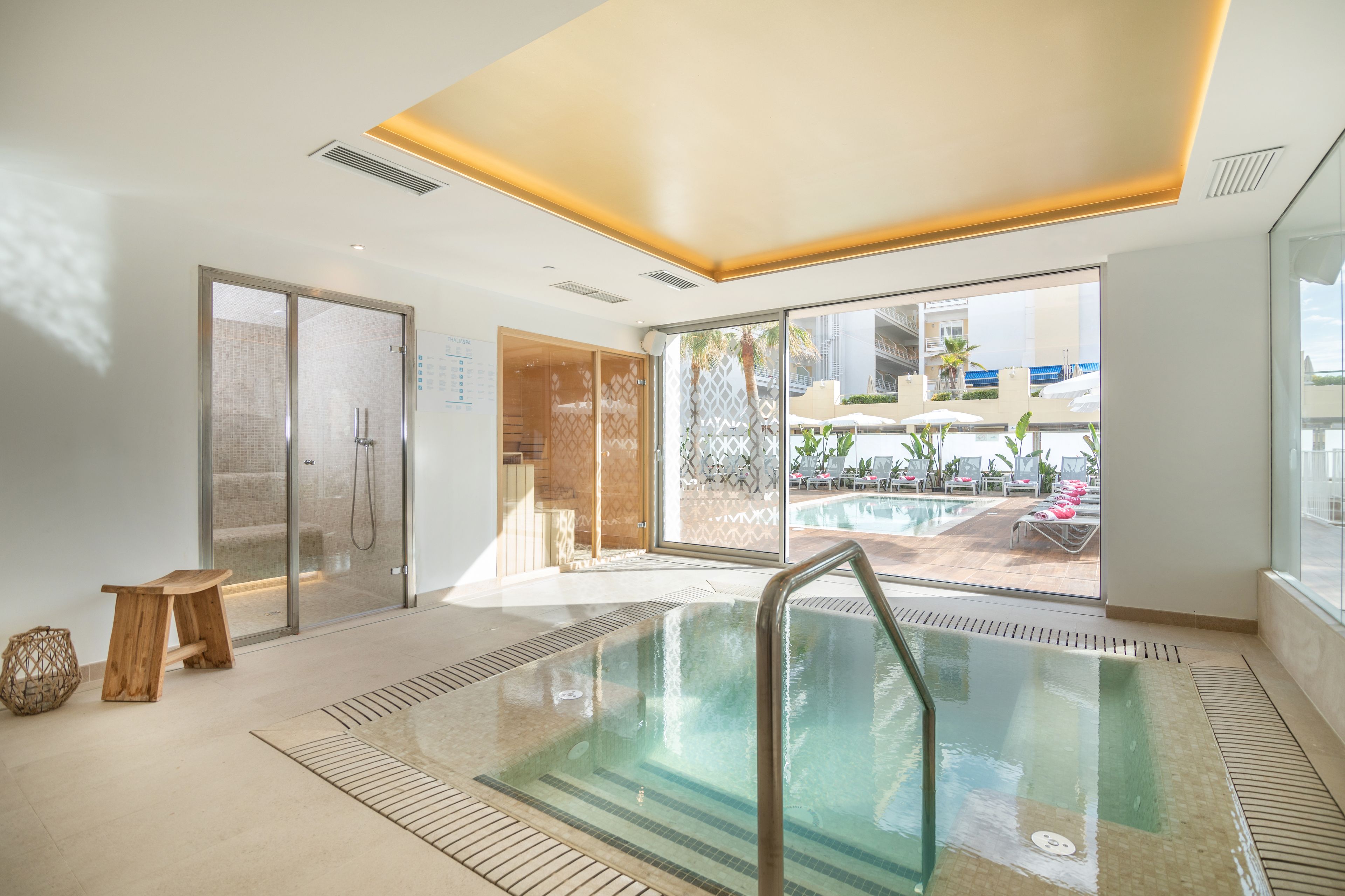 A modern indoor pool area with a glass door leading to a poolside area, a wooden bench, and a view of the outside.